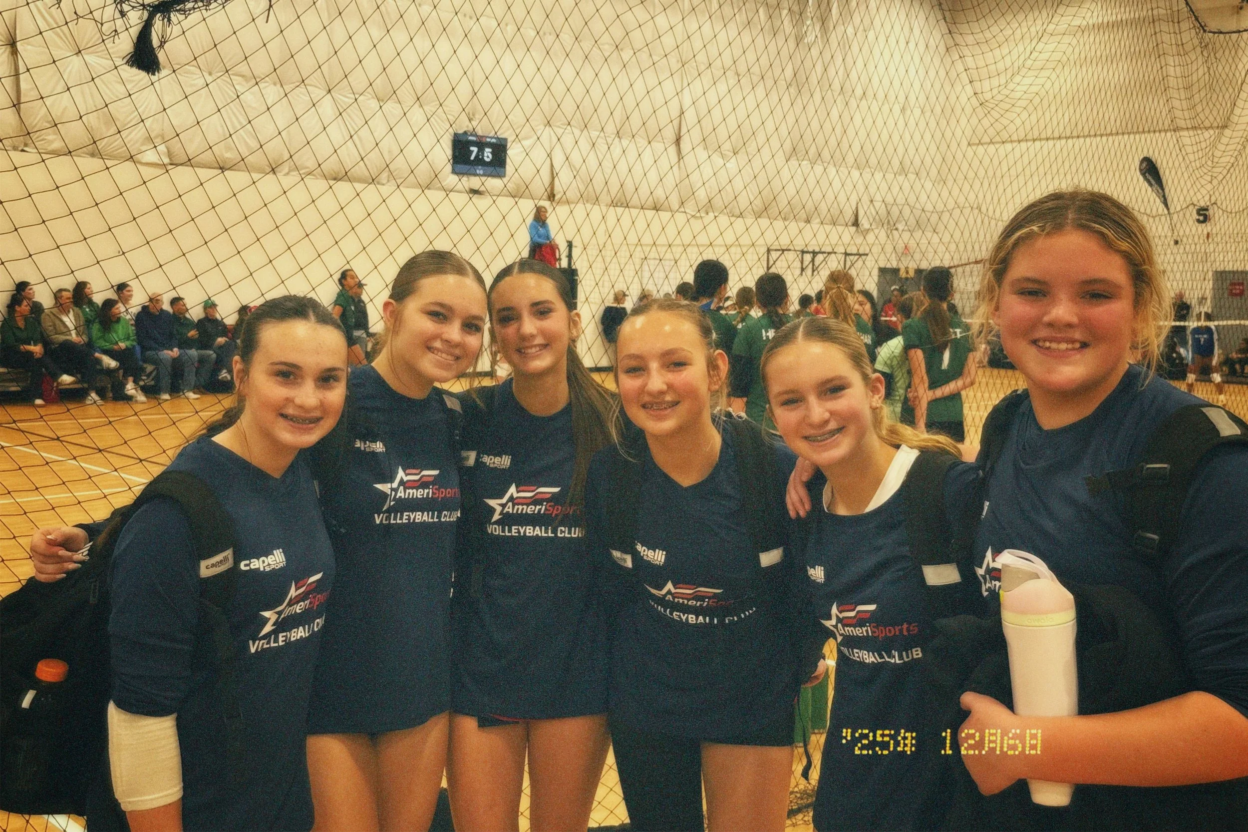 Six young female volleyball players standing together inside a gymnasium, wearing dark blue uniforms with logos, smiling at the camera. Behind them, an indoor volleyball court with a net, spectators sitting on benches, and a score display board.