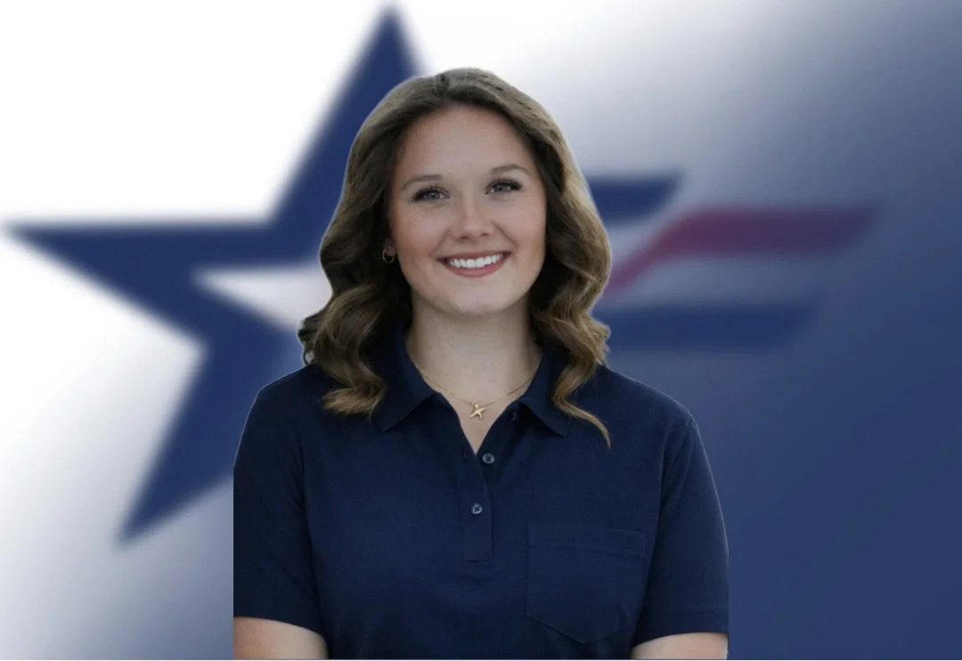 A woman with wavy, shoulder-length brown hair, smiling, wearing a dark blue collared shirt, standing in front of a blurred background with a large star symbol.