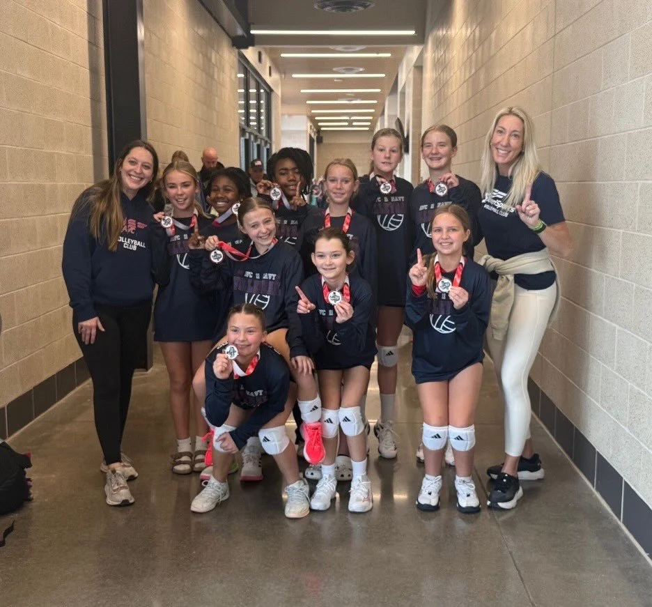 A group of young female volleyball players in matching navy blue uniforms and medals, posing with their smiling coaches in a hallway.