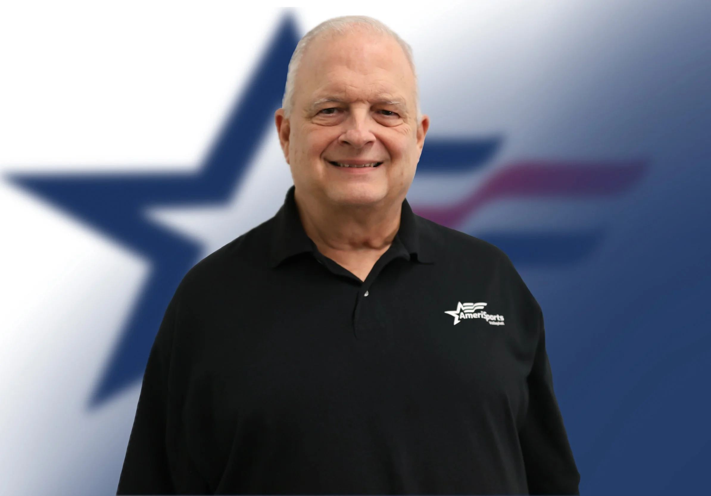 A smiling older man with short white hair wearing a black polo shirt with a logo that says "AmeriSports." He is standing in front of a blurry background featuring a large American flag and a star.