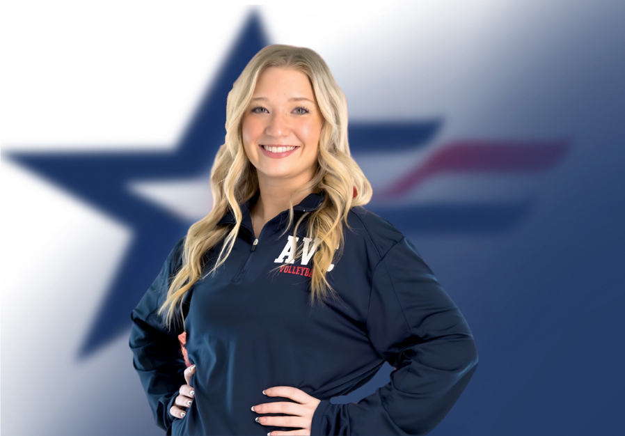A blonde woman in a navy blue volleyball jacket with 'AV' and 'VOLLEYBALL' on it, smiling, standing in front of a blurred background with a large star and American flag elements.