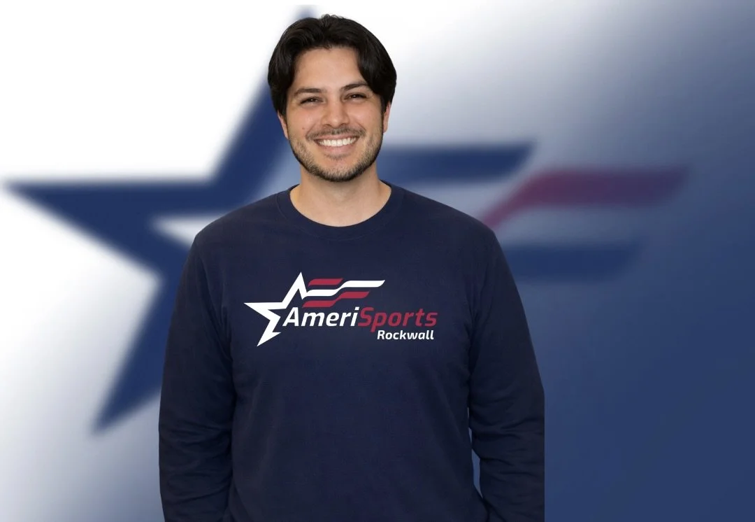 Man smiling wearing a navy blue AmeriSports Rockwall sweatshirt, standing in front of a blurred American Airlines logo background.