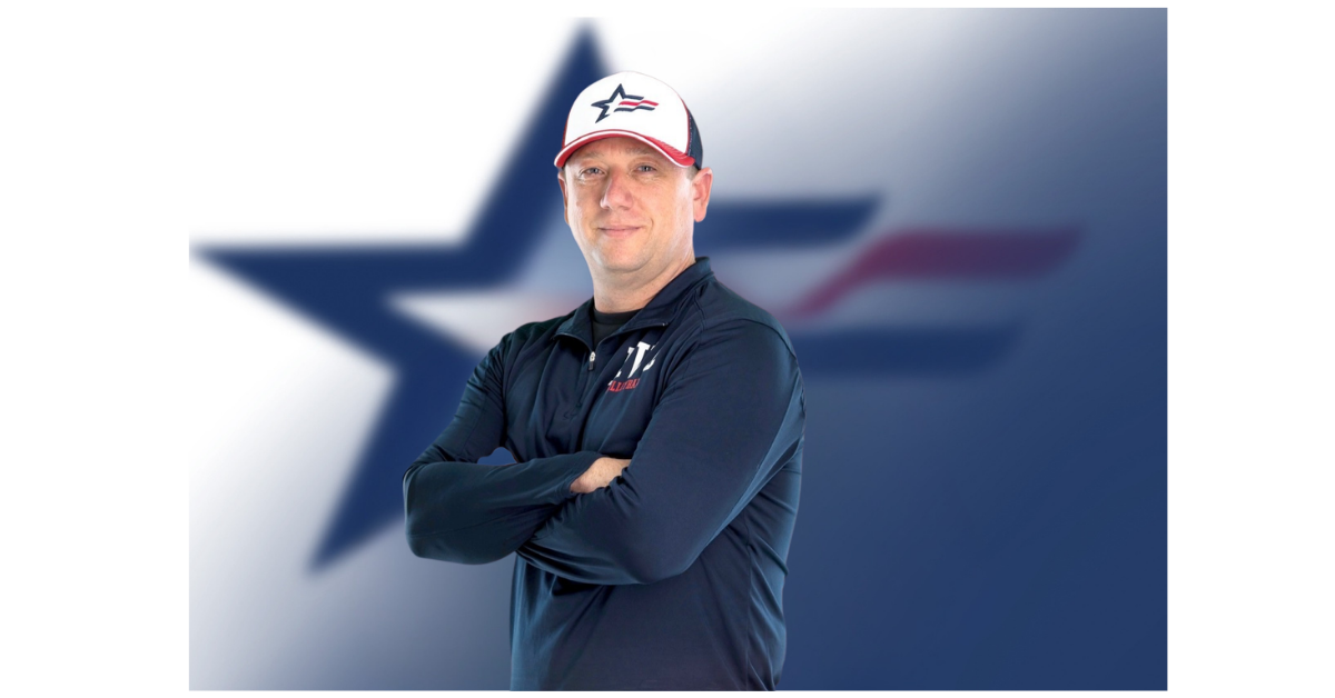 Man wearing a baseball cap and a navy polo shirt with logos, standing in front of a blurred Texas flag background.