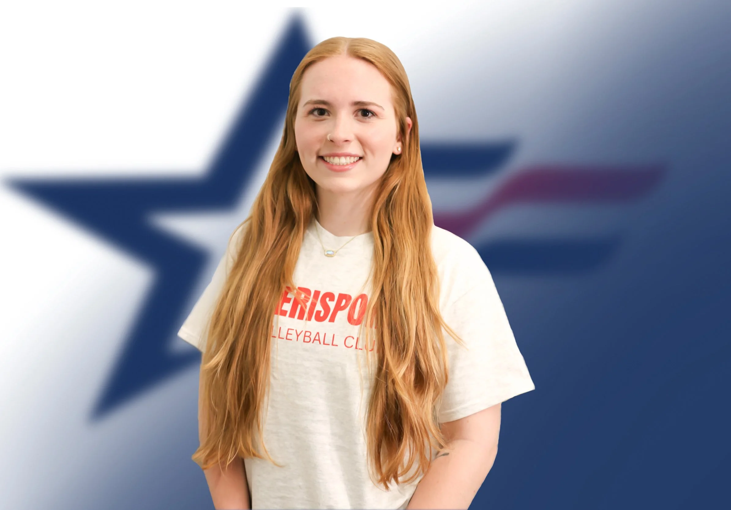 A young woman with long red hair, smiling, wearing a white t-shirt with red text, standing in front of a blurred background featuring a blue star.