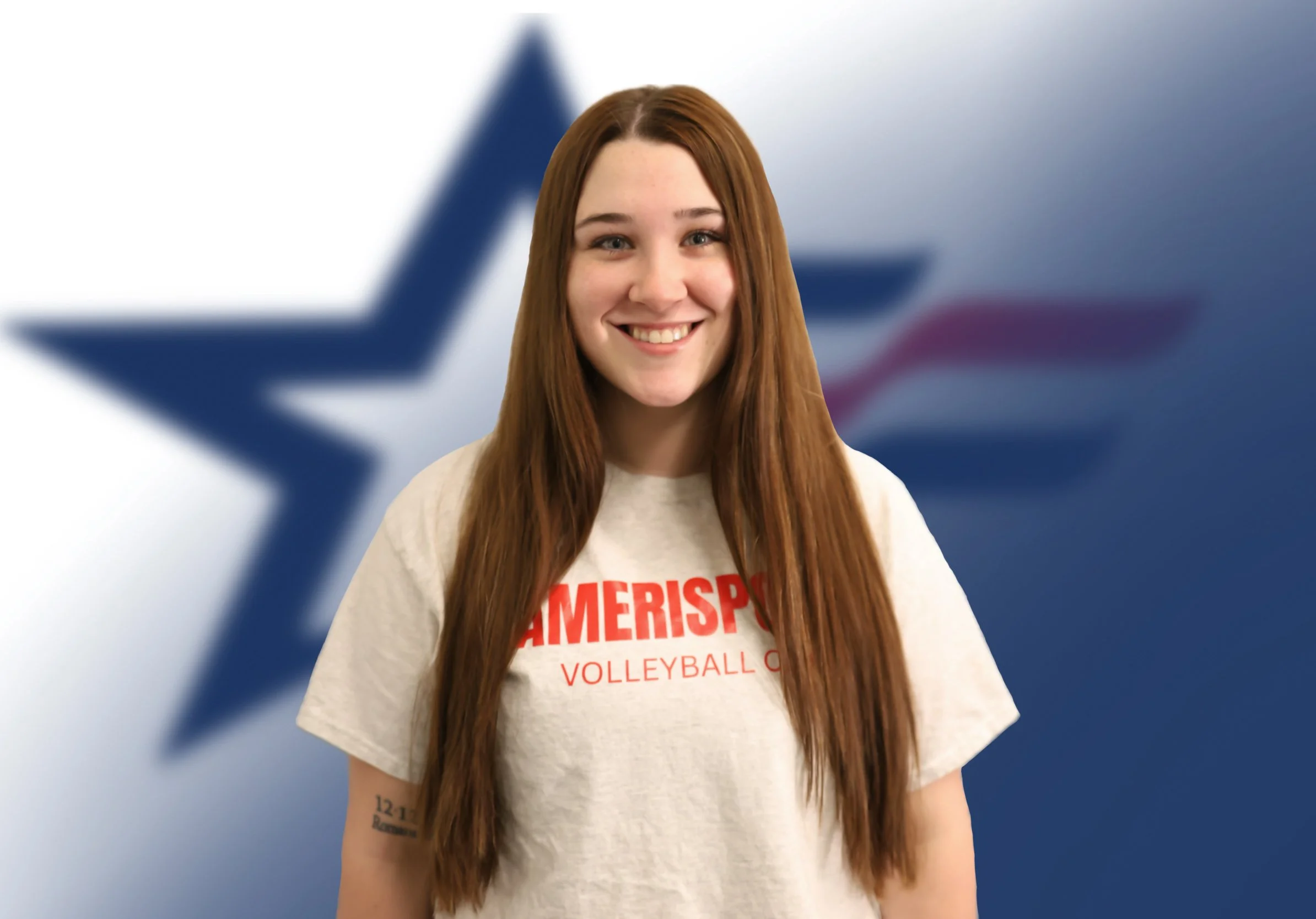 Young woman with long red hair smiling, wearing a cream shirt with red text, standing in front of a blue background with a stylized American flag in the background.
