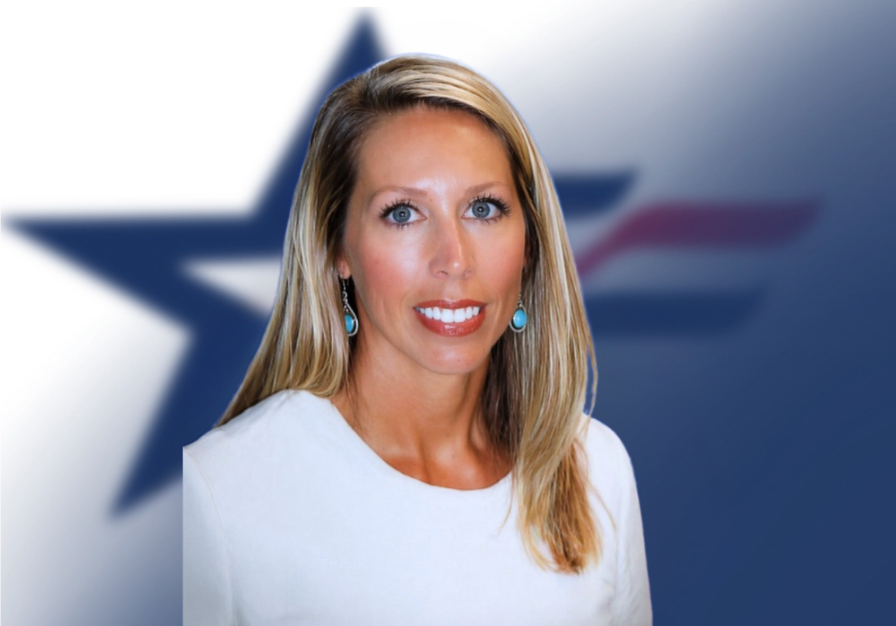 A woman with blonde hair, blue earrings, and a white top standing in front of a blurred American flag background.