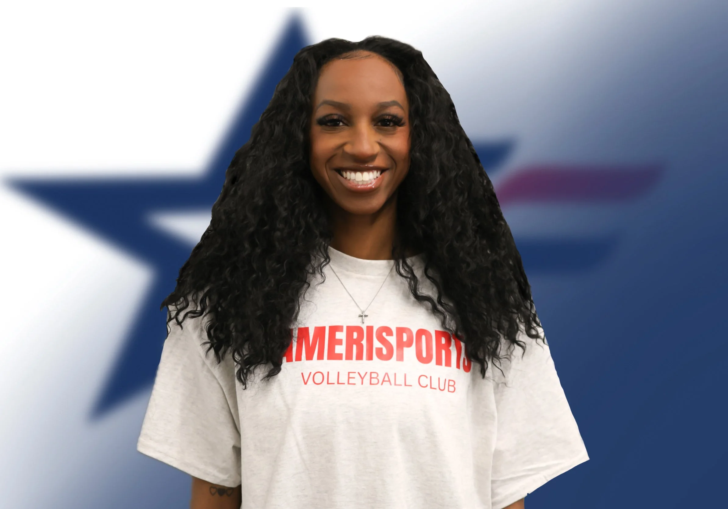 A woman with curly hair smiling, wearing a white shirt with red text that says 'GAMERSPORT VOLLEYBALL CLUB' and a small cross necklace, standing in front of a blurred background with a blue and white star.
