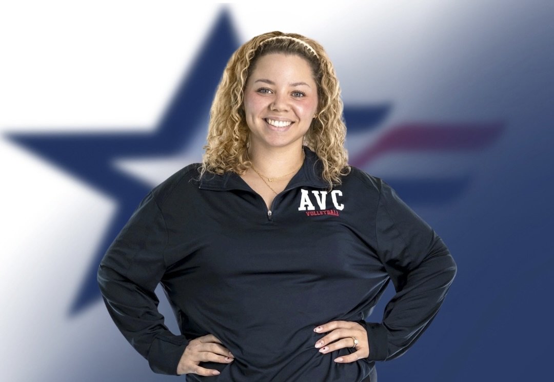 Young woman with curly blonde hair smiling, wearing a navy volleyball jacket with 'AVC' and 'Volleyball' embroidered, standing in front of a blurred American flag background.