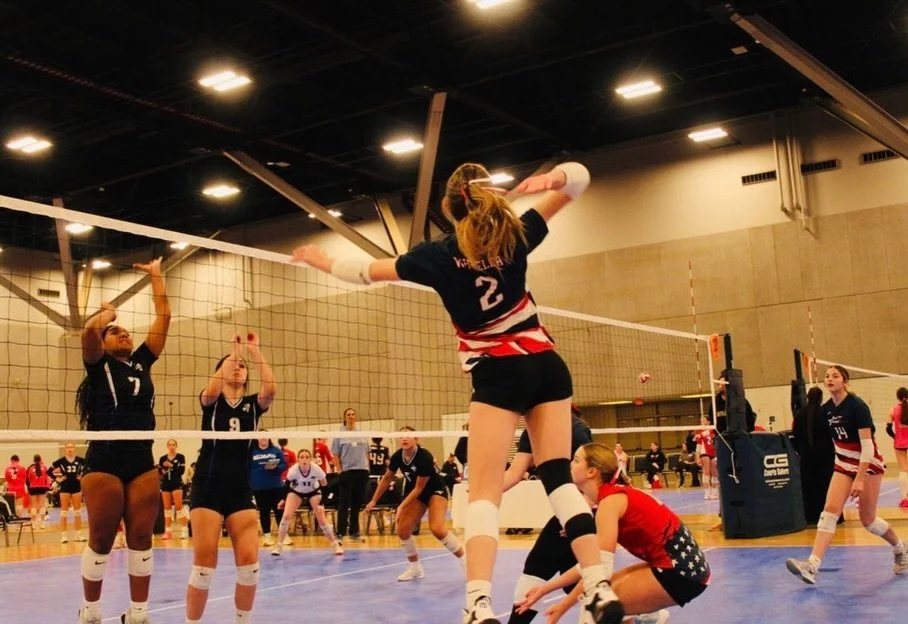 Women's volleyball game in an indoor gym, with players jumping to block or spike, and others preparing on the court.