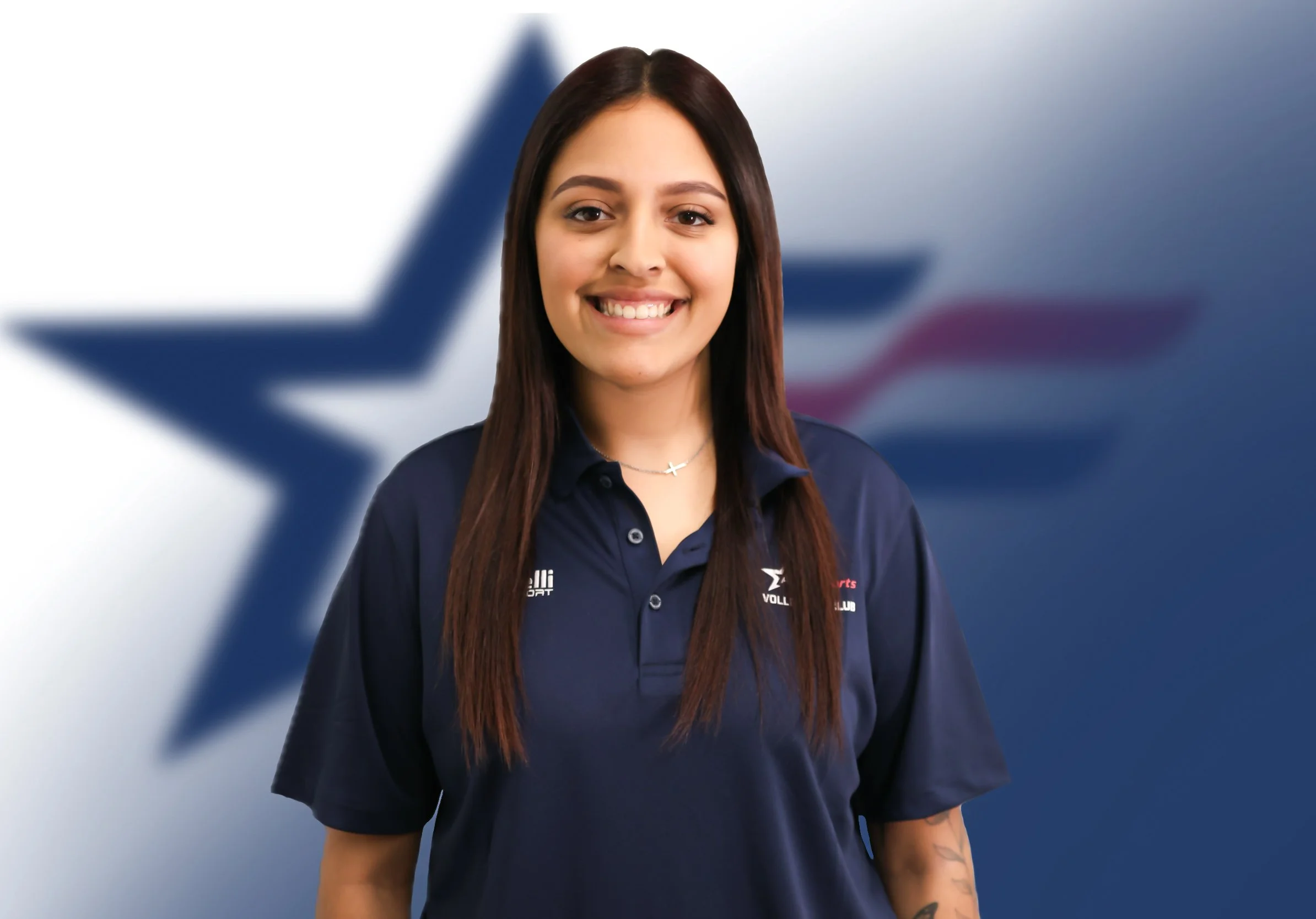 A young woman in a dark blue uniform standing in front of a blurred background featuring a star logo. She is smiling and has long dark hair and a small cross necklace.