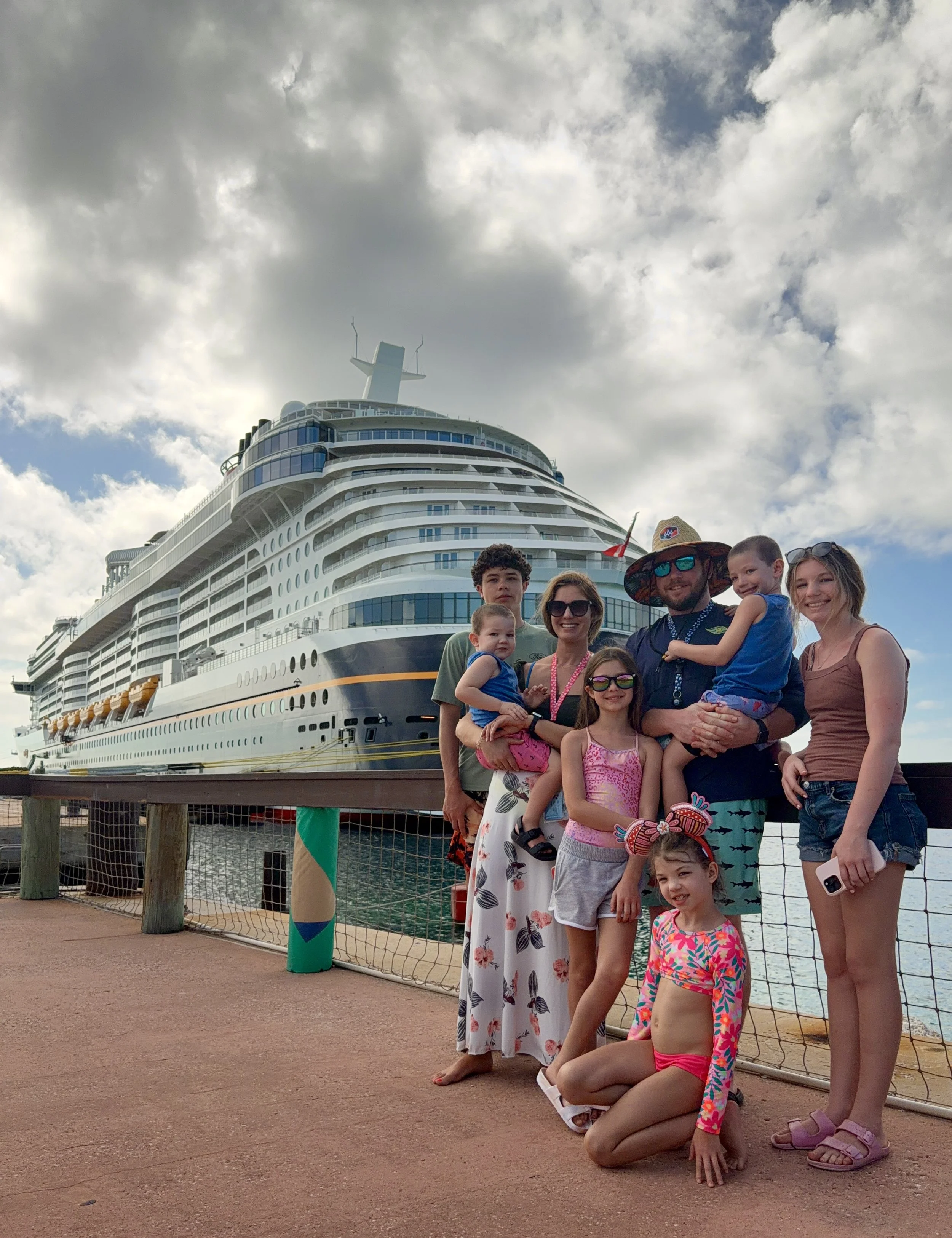 A group of smiling family members, including children and adults, are standing together near a ship dock, with cloudy sky overhead. The ship is large, white, and appears to be a cruise liner.