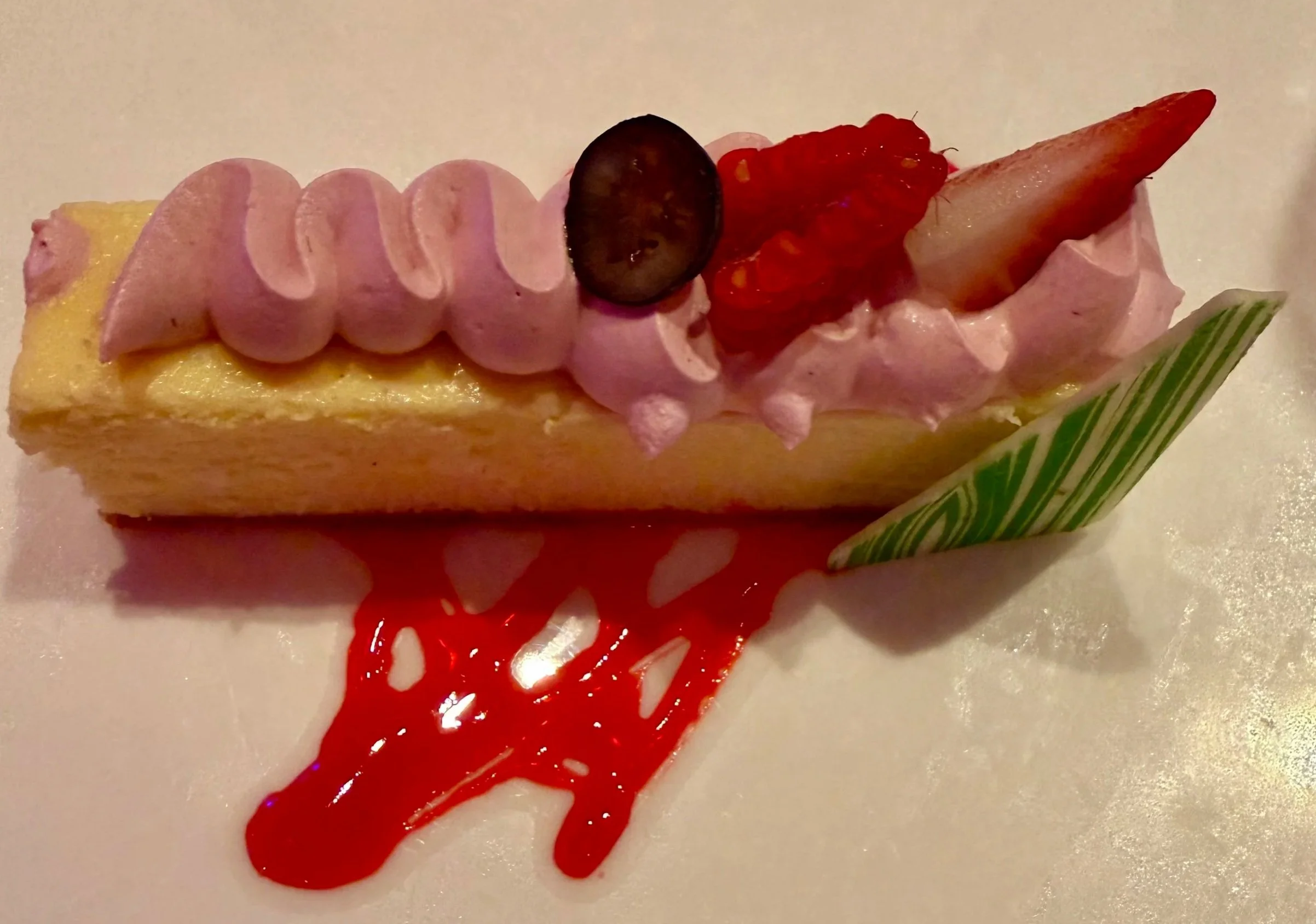 A dessert with a square yellow base, topped with pink whipped cream, decorated with slices of pink fruit, a dark purple grape, a red fruit topping, and a small green-and-white striped piece.