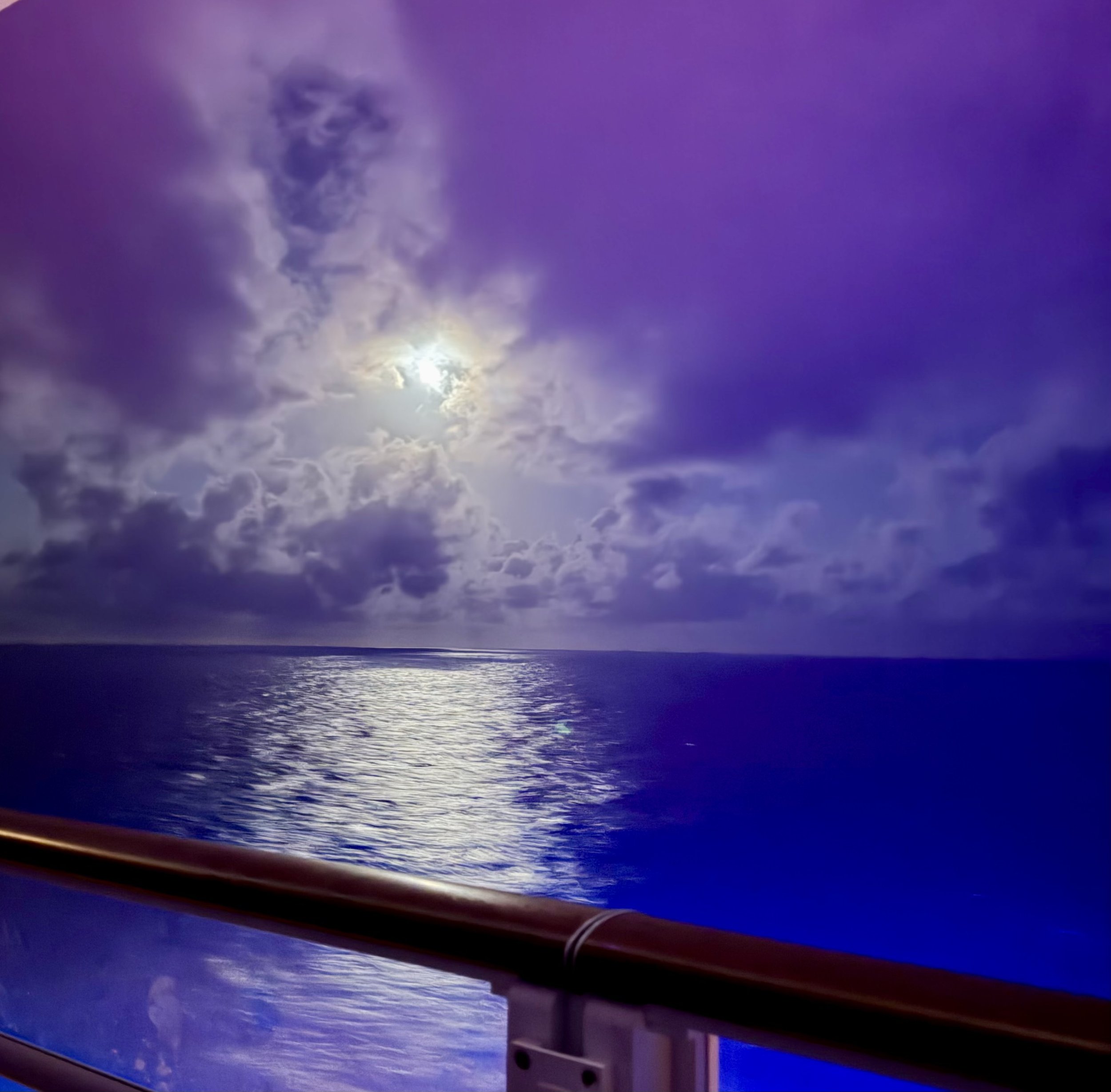 A view over the ocean at night with moonlight reflecting on the water. The sky is partly cloudy with a large, bright moon and some clouds around it.