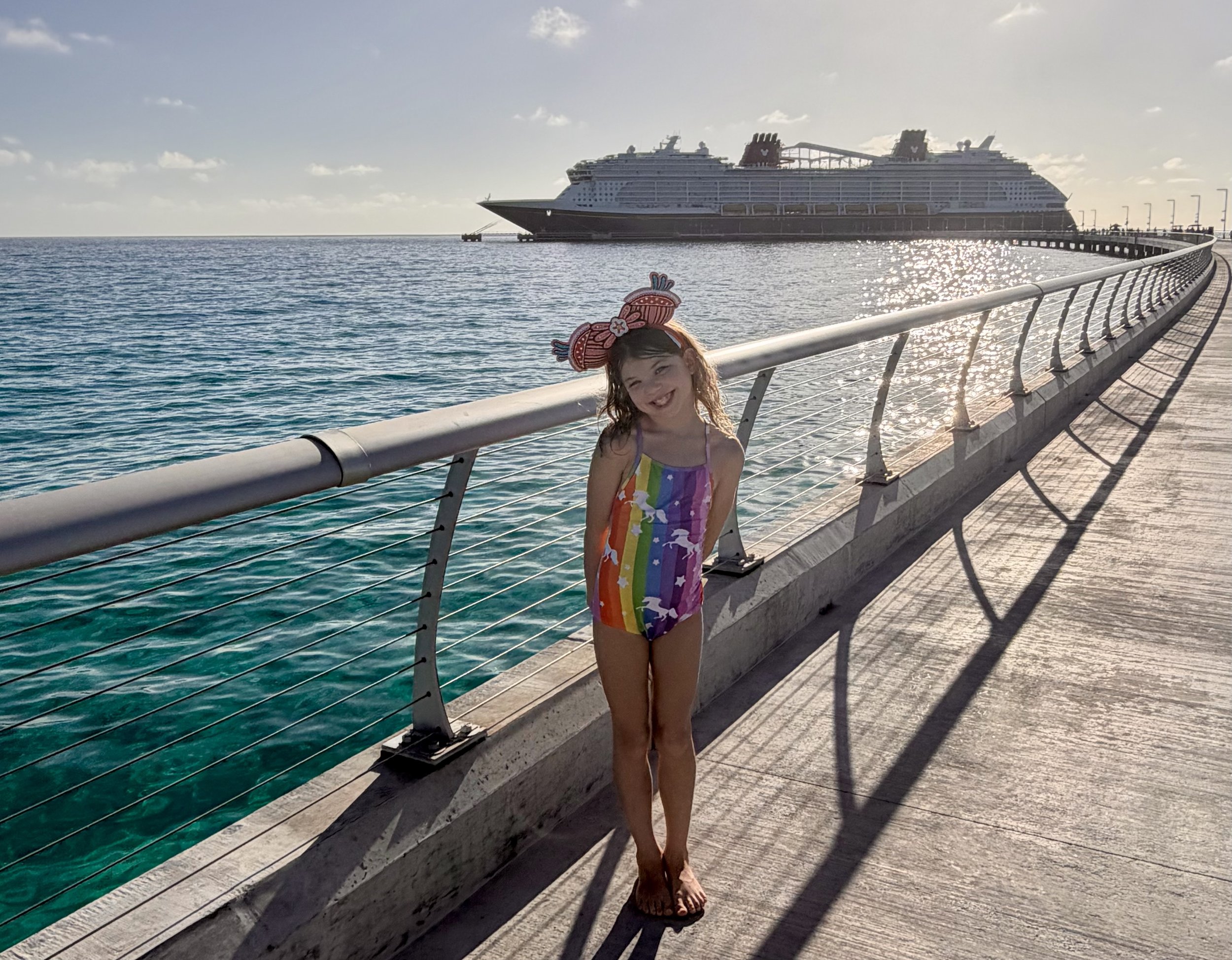 A young girl with red hair and a rainbow-colored dress standing on a pier, smiling, with a large cruise ship in the background, the ocean, and a partly cloudy sky.