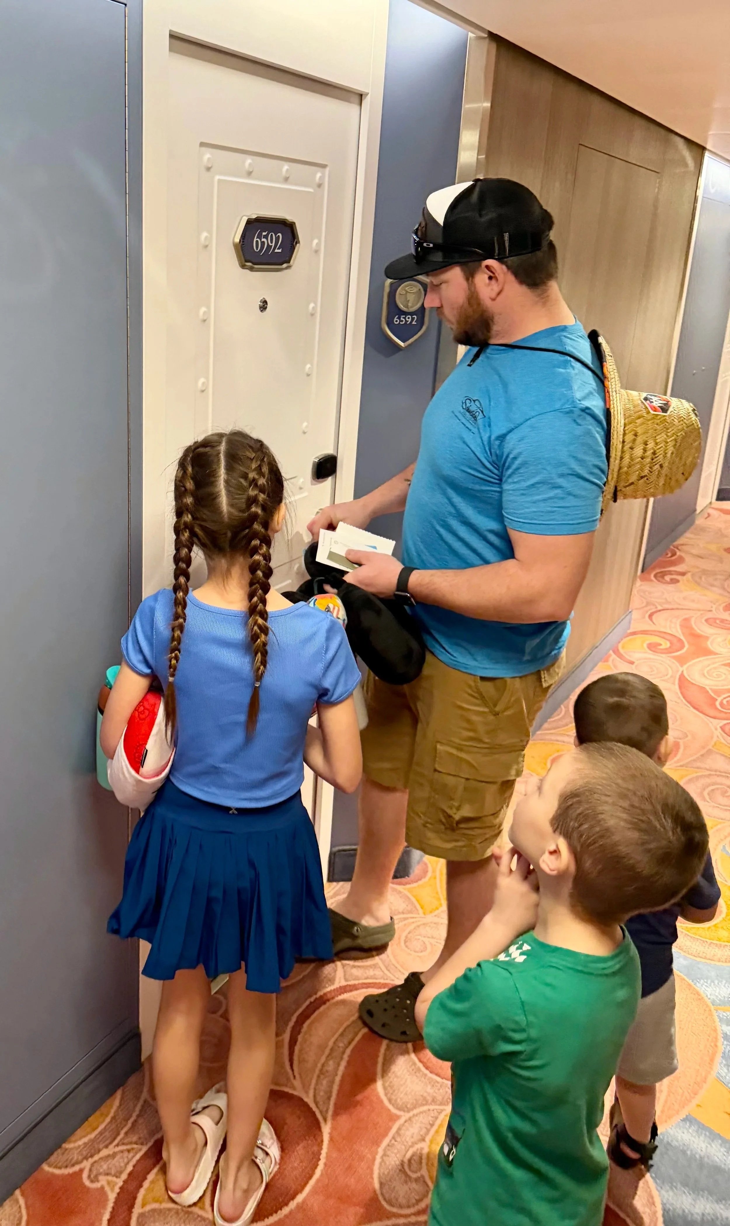 A group of children and an adult standing in line outside a stateroom, with room number 6592, in a hallway with colorful patterned carpet.