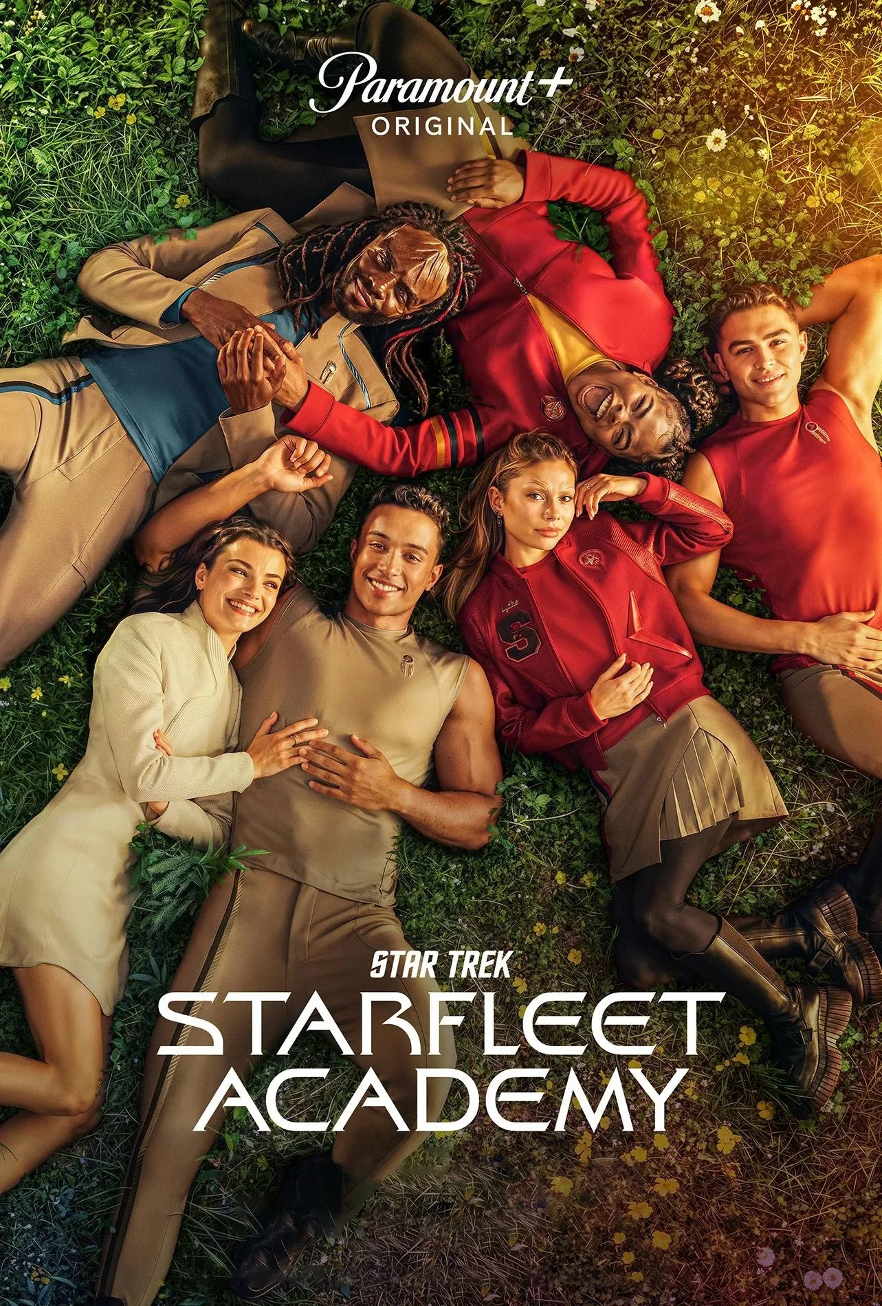 Six diverse people lying on grass with small yellow and white flowers, smiling and relaxed. They are wearing casual and athletic clothing, with some in red, tan, and beige outfits. The scene is bright and cheerful, with the text "Paramount+ Original" at the top and "Star Trek: Starfleet Academy" at the bottom.