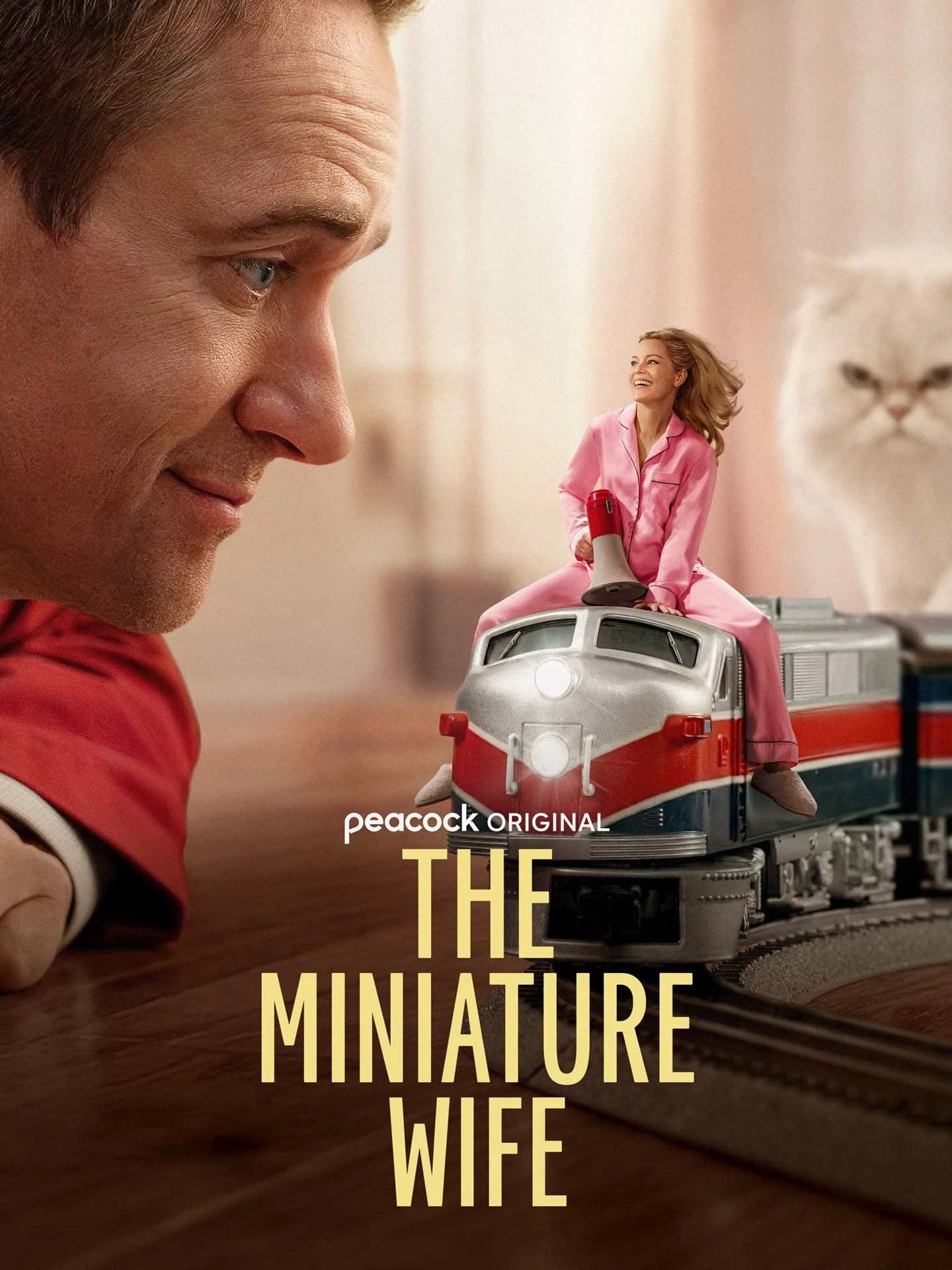 Close-up of a smiling woman in pink pajamas riding a miniature train. In the foreground, a man is looking down, with part of his face visible. The background shows a blurry large white cat statue and soft pink walls. The text reads "peacock ORIGINAL THE MINIATURE LIFE WIFE."