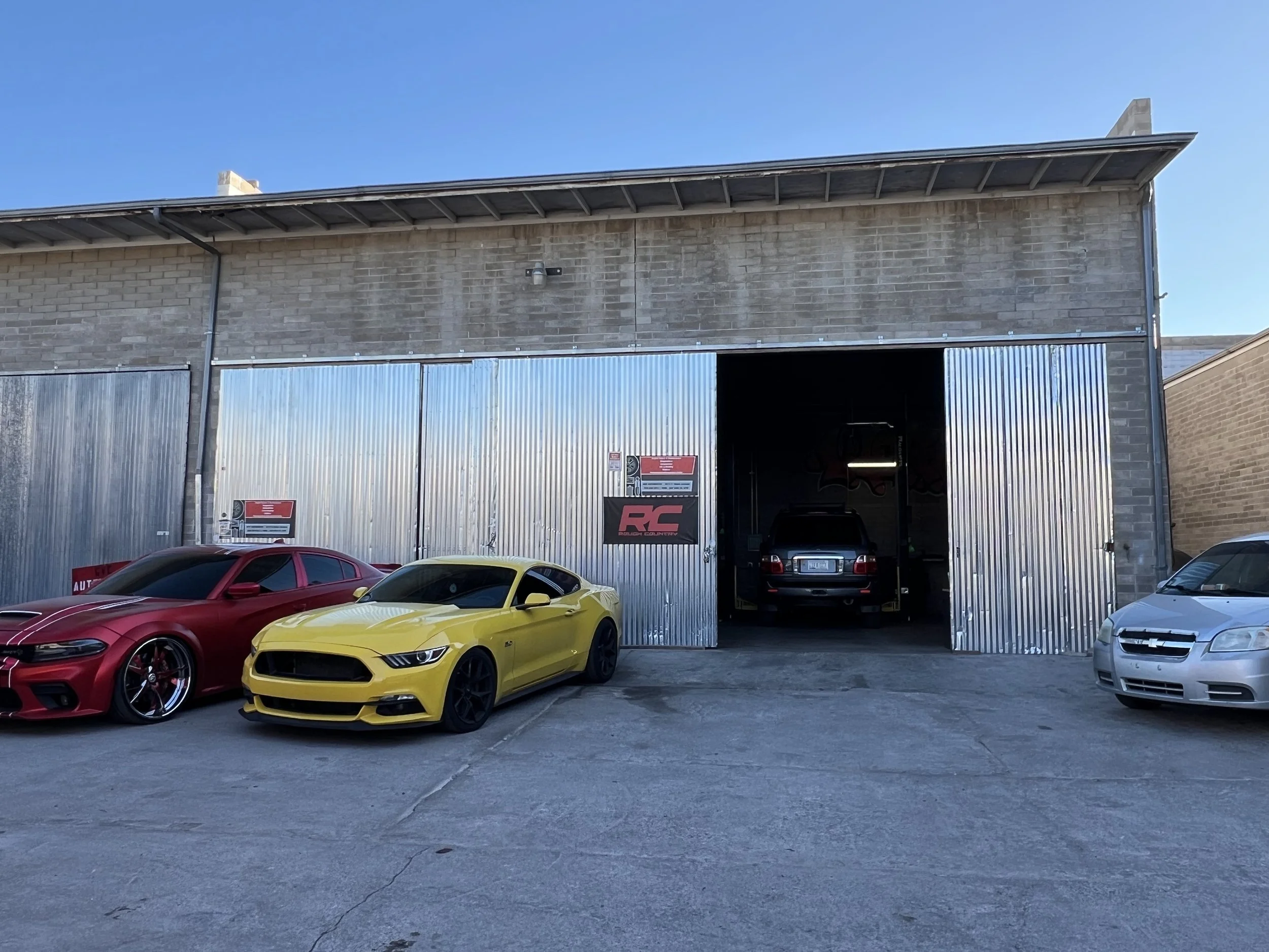 A building with a brick upper section and a large open garage door, partially covered with corrugated metal sheets, under a clear blue sky. In front of the building, there are several sports cars, including a red car, a yellow car, and a white car, parked on a concrete surface.