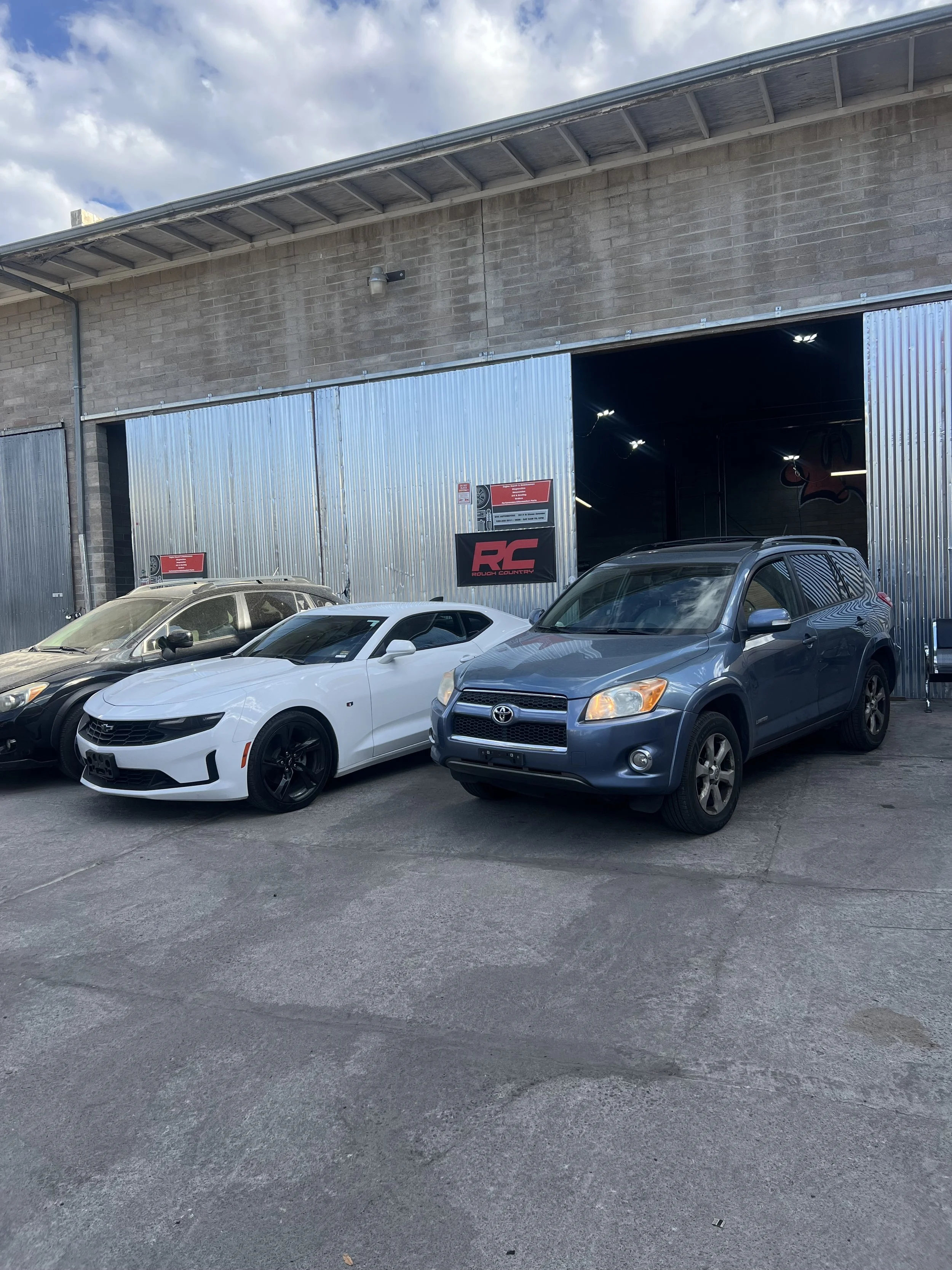 A parking lot with four cars parked in front of a large industrial building with metal sliding doors and a brick wall. The sky is partly cloudy.