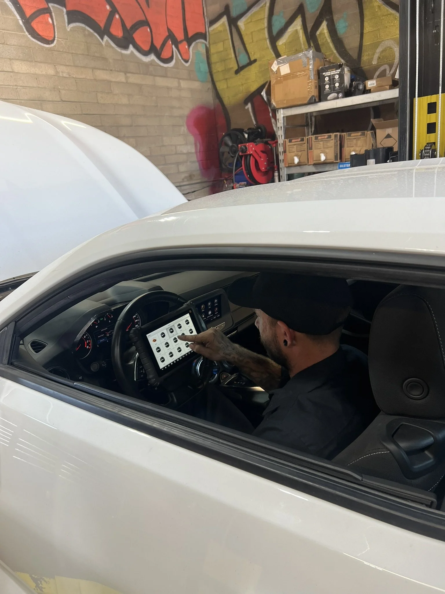 A man with a beard and black hat sitting in the driver's seat of a white vehicle, using a tablet or smartphone. The vehicle's window is rolled down, revealing an indoor setting with a brick wall that has graffiti art, and a shelf with boxes and equipment, including a red and black tool or device.