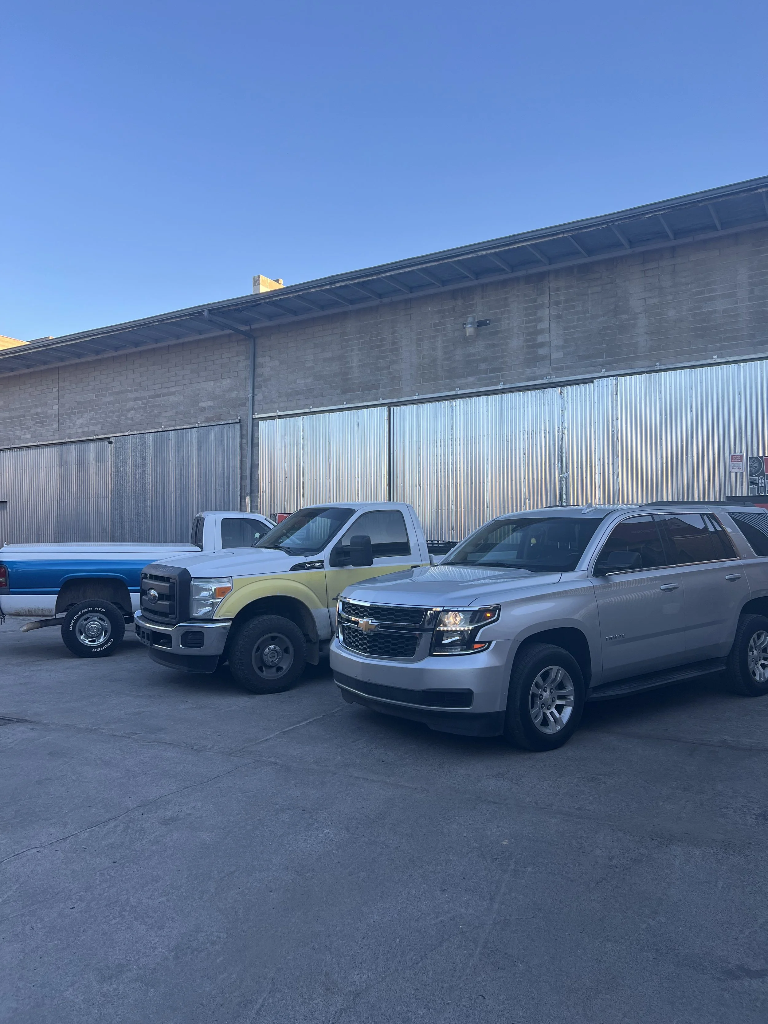 A parking lot with four parked vehicles, including a silver SUV, a white pickup truck, and two trucks, in front of a large brick building with a corrugated metal fence and a clear blue sky.