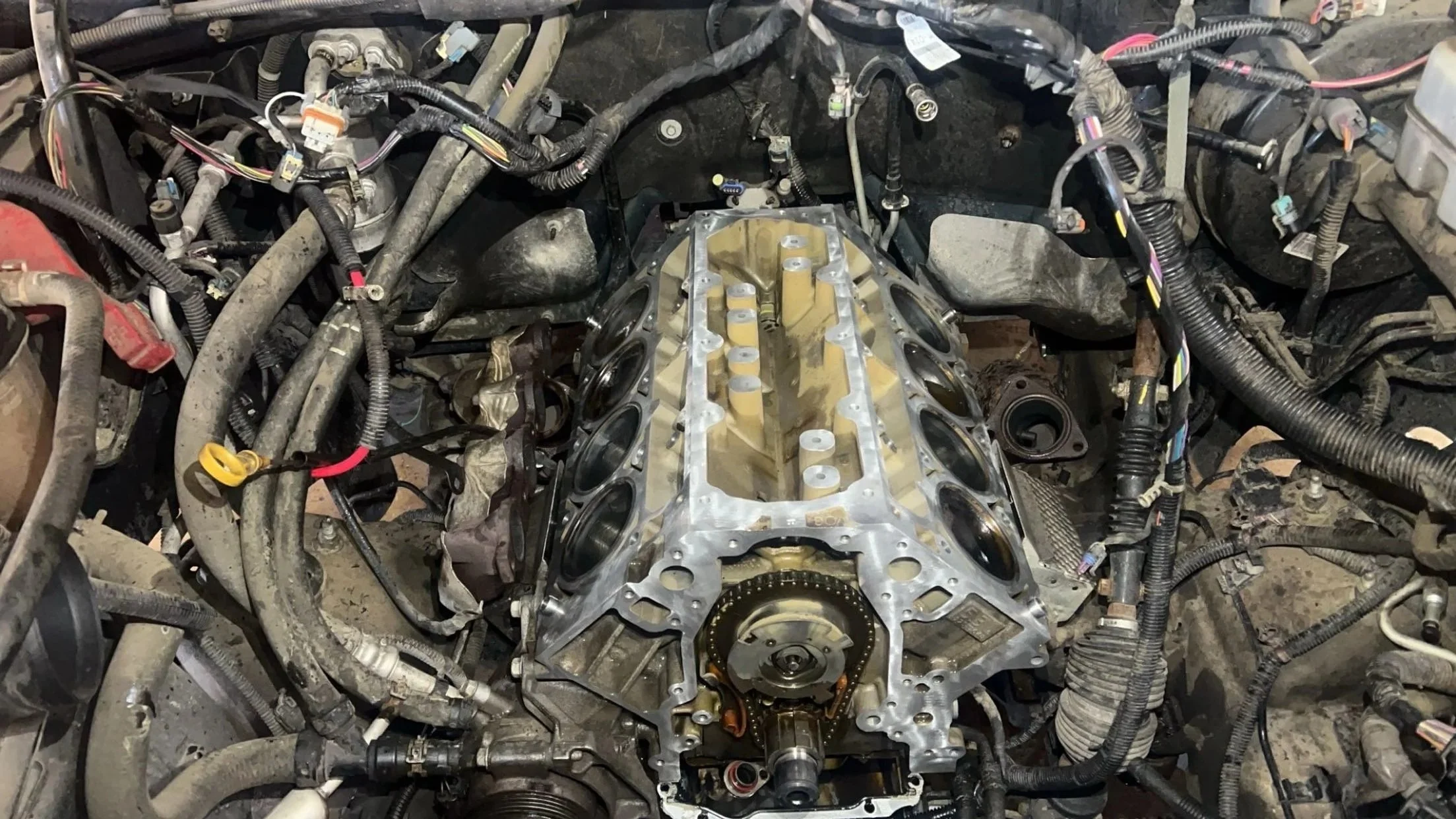 View of a car engine bay with the engine removed, showing the engine block, timing chain, and various wiring and hoses surrounding it.