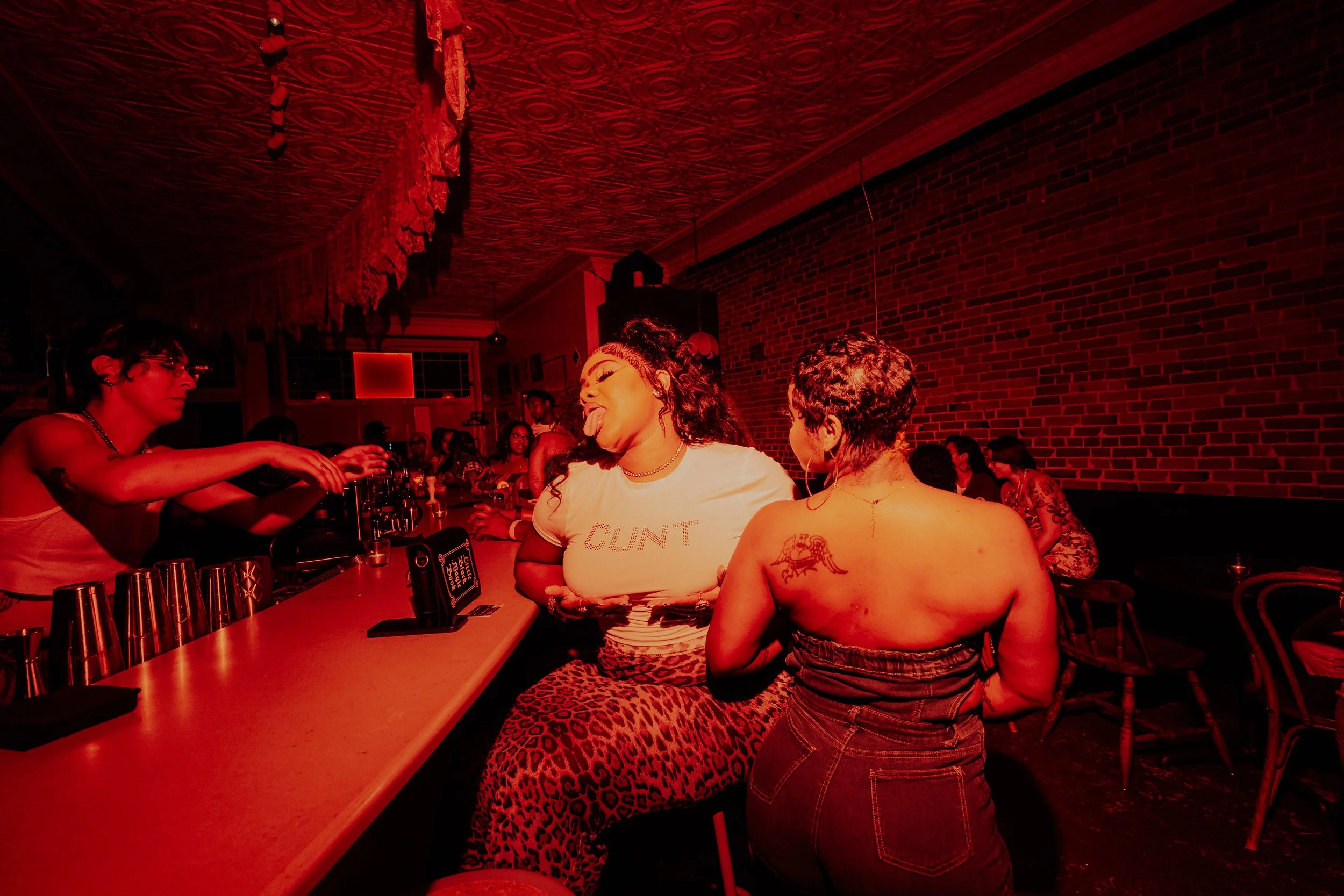 A scene inside a dimly lit bar with a warm red hue. Several women are gathered around a bar counter, some with tattoos and leopard print clothing, enjoying drinks and socializing. One woman is sticking her tongue out playfully, and a bartender is reaching out to her. The background features a brick wall and a ceiling with ornate detailing.