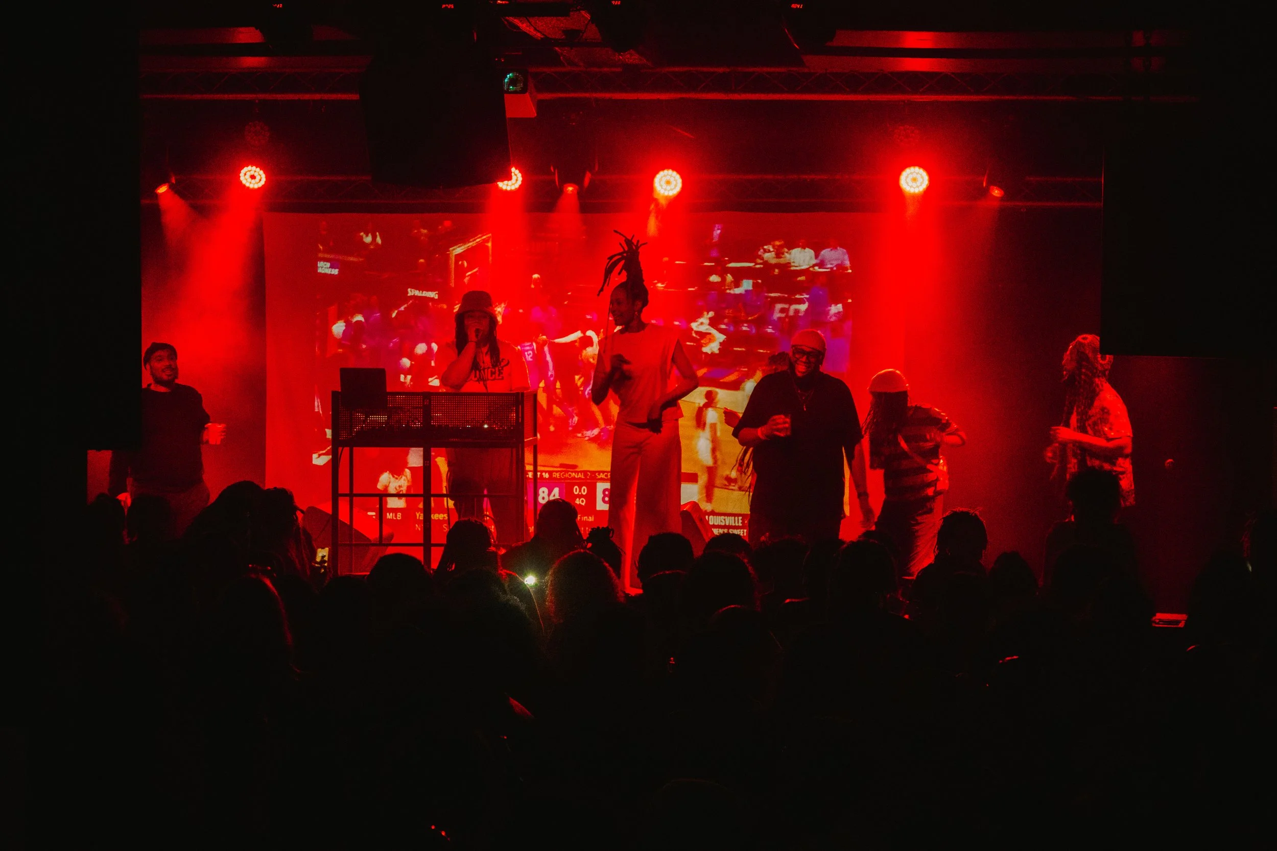 People standing on a stage with red lighting, some speaking into microphones, in front of a large screen displaying a sports game, against a dark background with an audience visible.