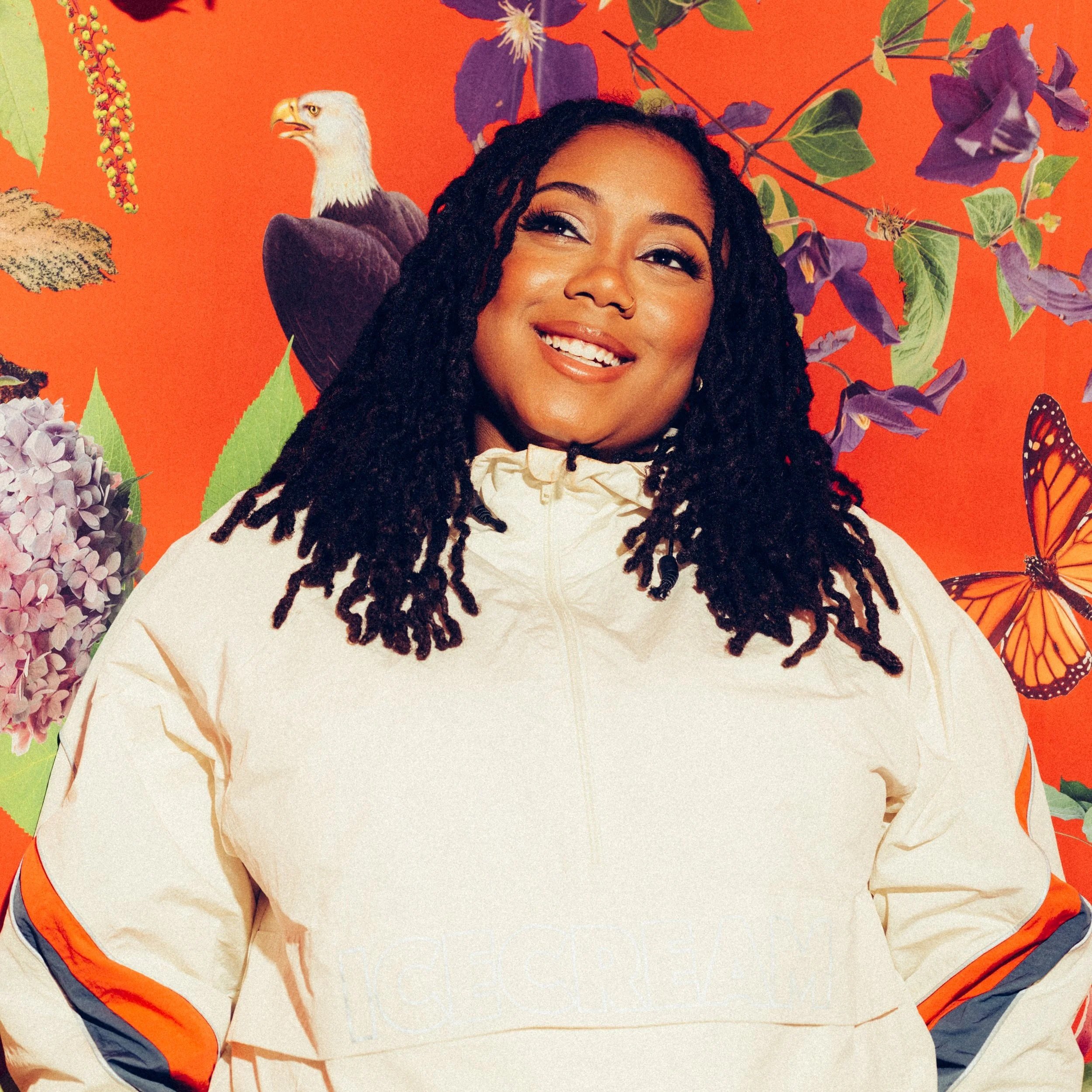 A smiling woman with long locs poses against a vibrant orange background decorated with butterflies, flowers, and an eagle