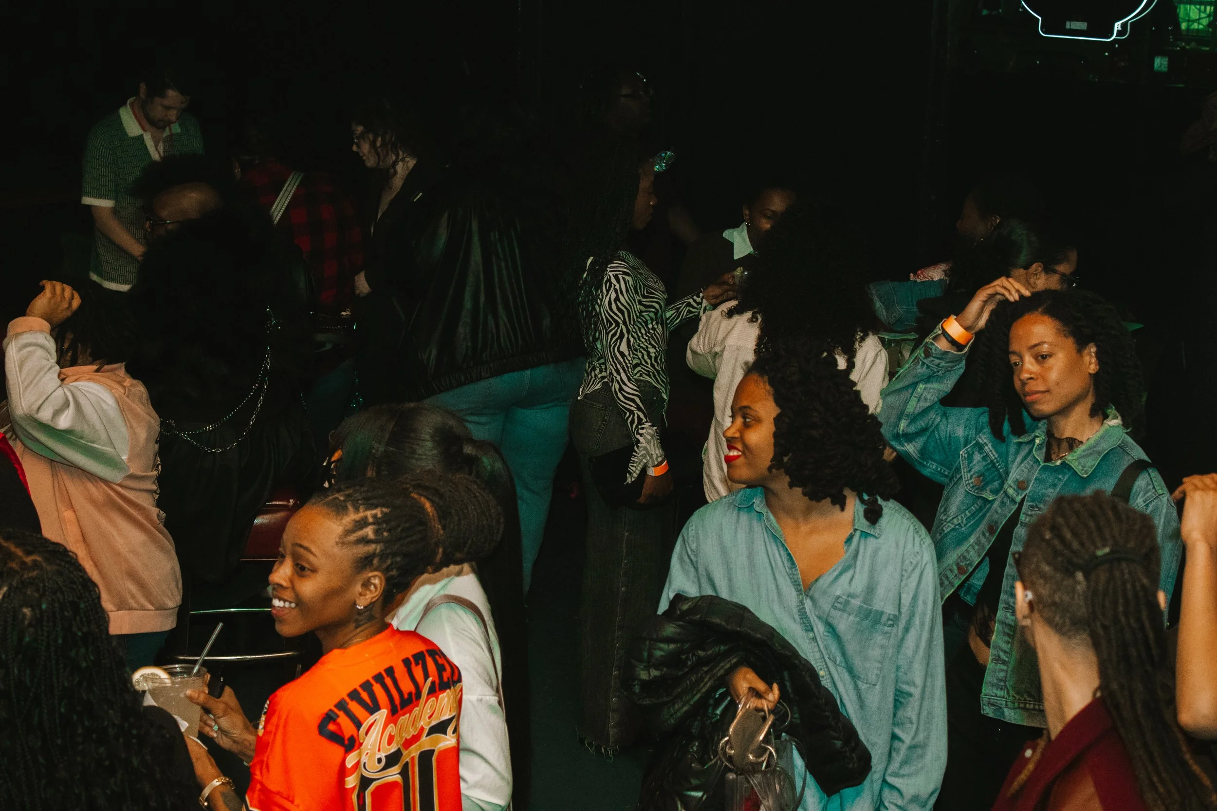 A group of people gathered together in a crowded indoor setting, some are standing and talking, others are sitting, with dark background and neon lights.