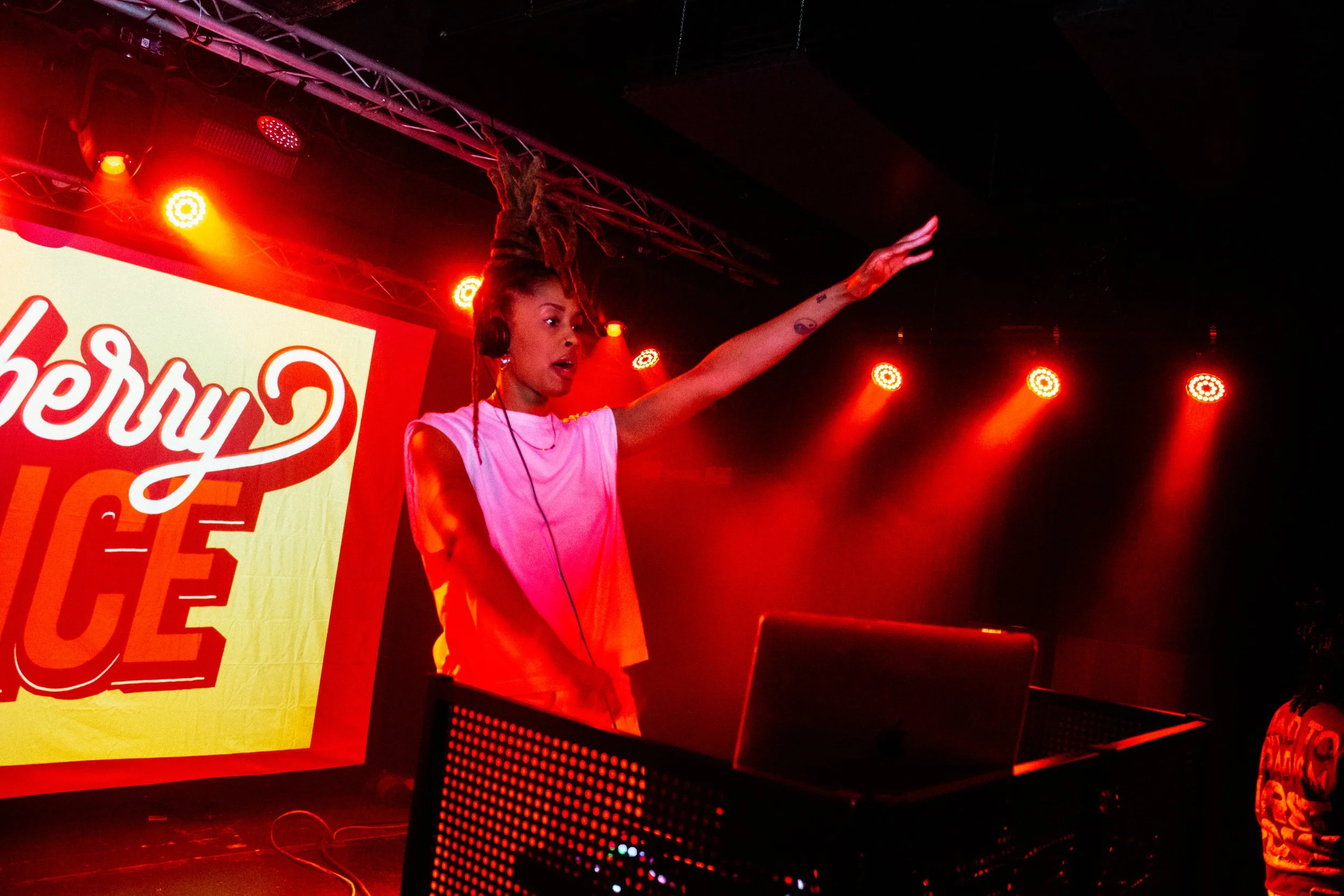 A woman with long braided hair and tattoos on her arm is DJing on stage, wearing headphones and a white sleeveless top, under red stage lighting, with a large bright sign partially visible in the background.