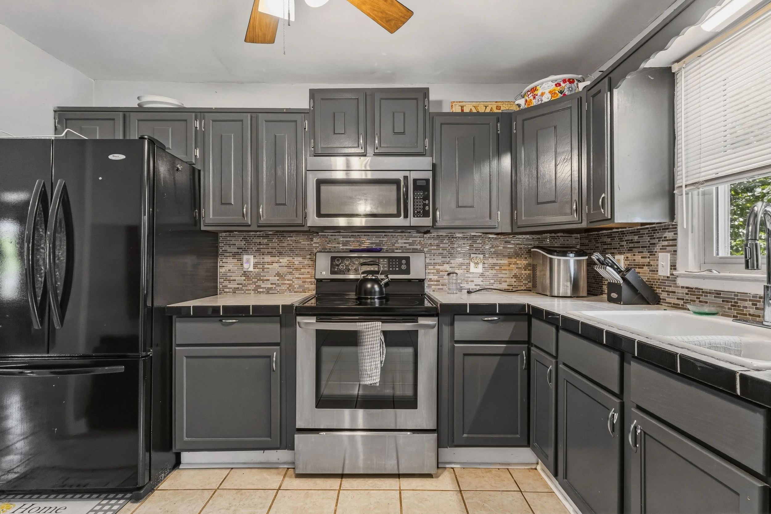 A kitchen with gray cabinets, black appliances, a mosaic tile backsplash, a microwave above the stove, a stainless steel dishwasher, a white countertop with a sink, window blinds, and various kitchen items on the counter.