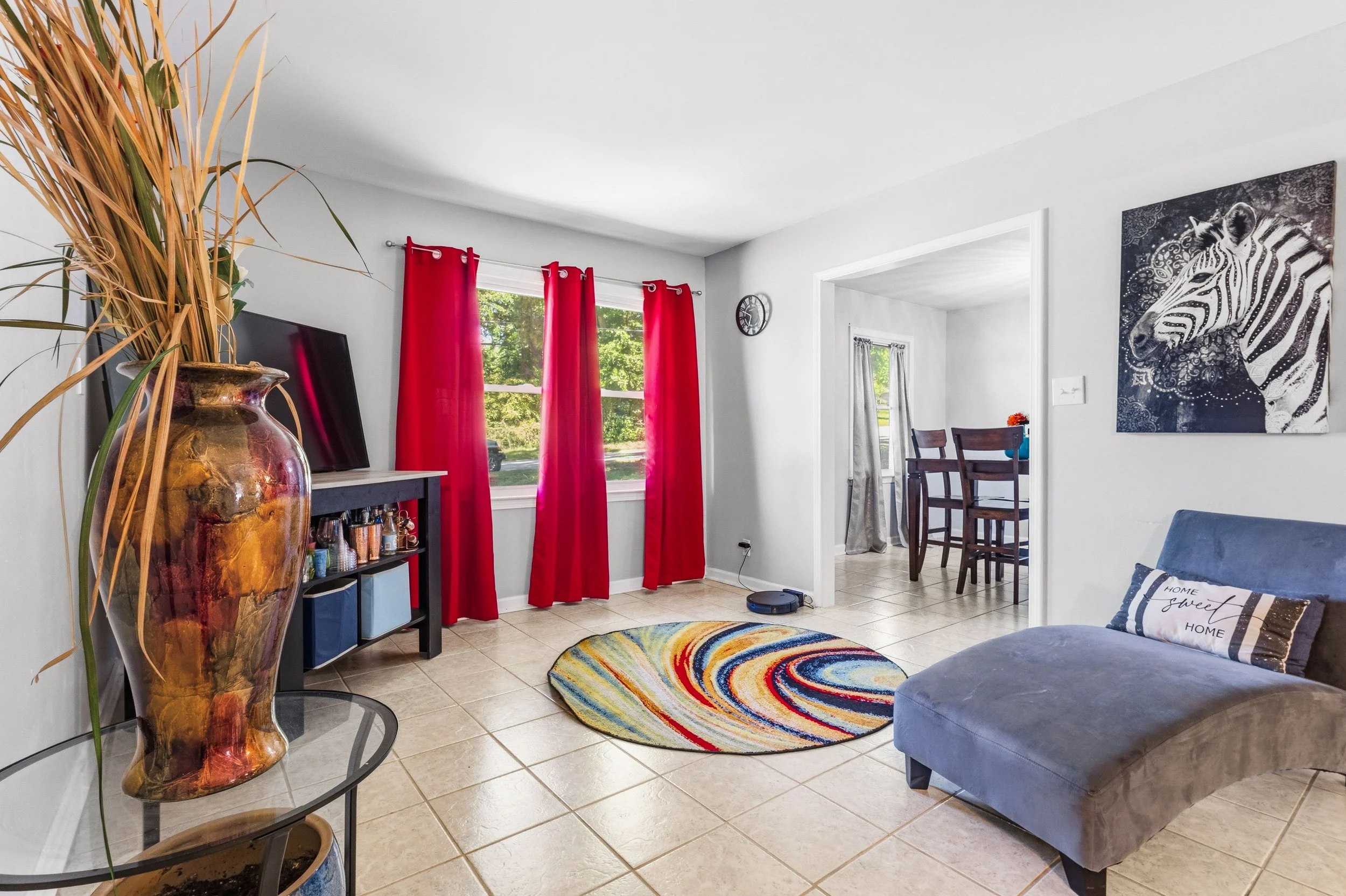 Living room with large window with red curtains, a colorful oval rug, a blue sofa with a decorative pillow, a black TV stand, a wall clock, and a zebra artwork on the wall.