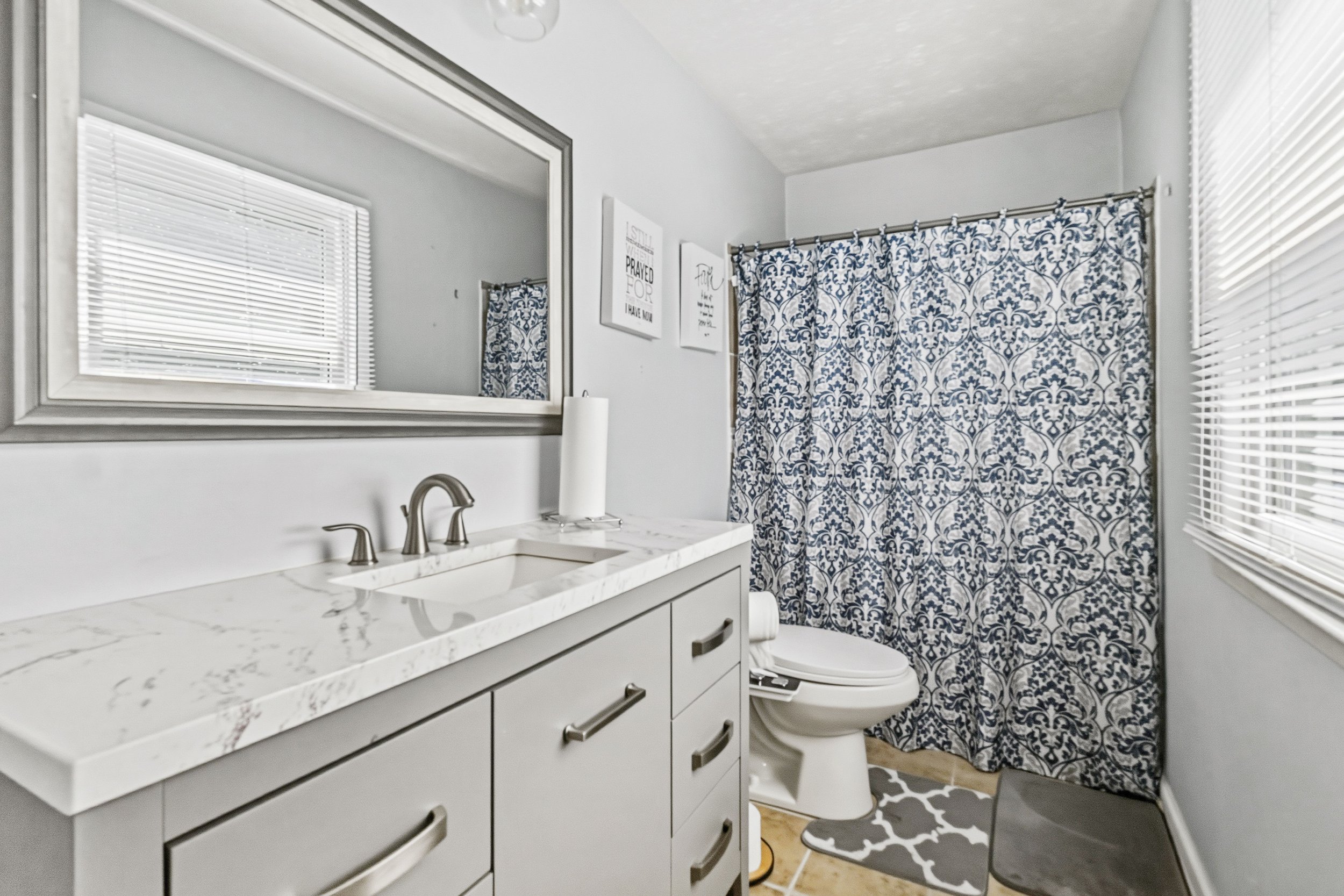 Bathroom with white vanity and marble countertop, large mirror, window with blinds, patterned shower curtain, and toilet with patterned rug.
