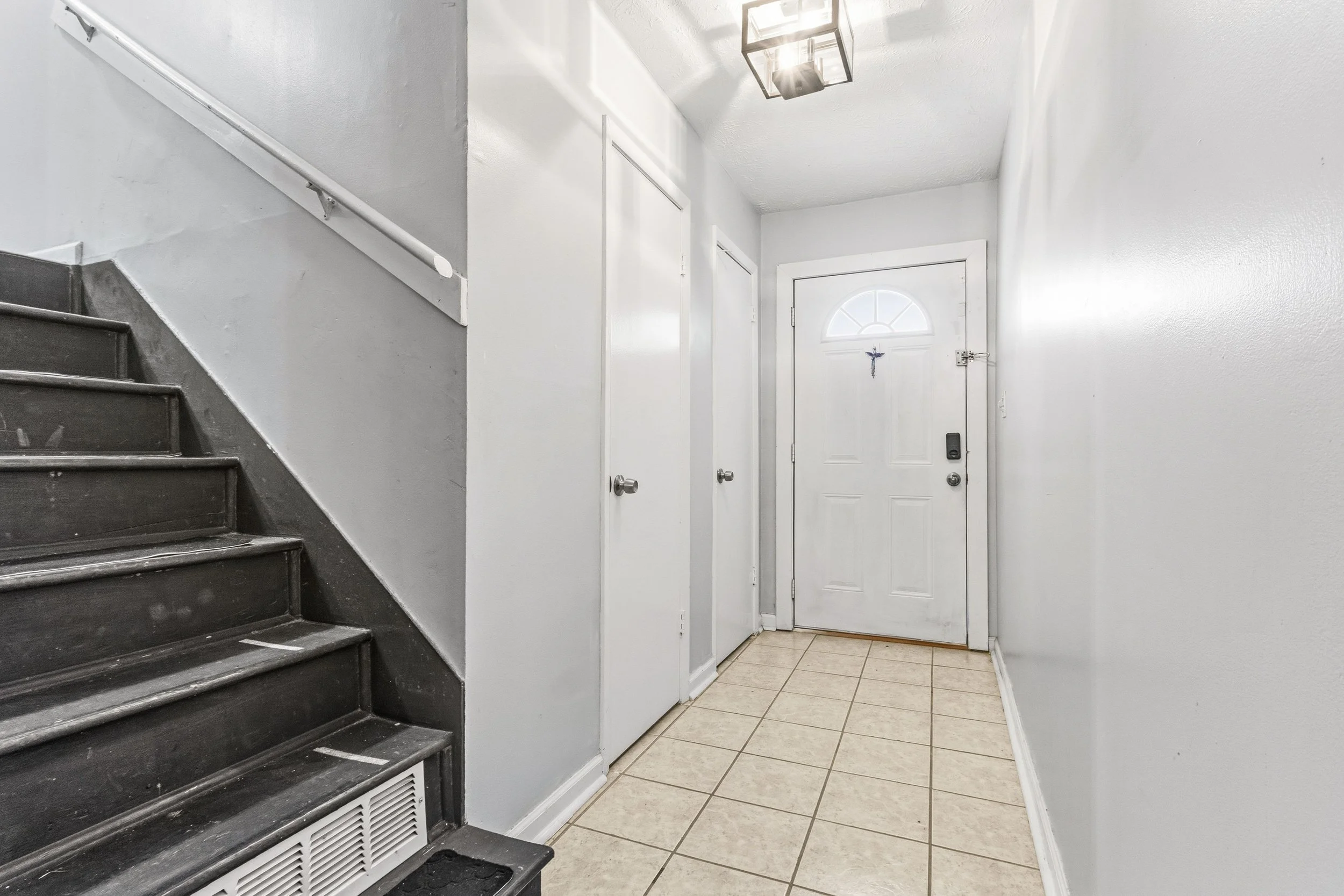 A small entryway with beige tile flooring, white walls, and a white door with a small arched window. To the left, there is a staircase with dark wooden steps and a white handrail. There are two closed closet doors or storage compartments on the wall.