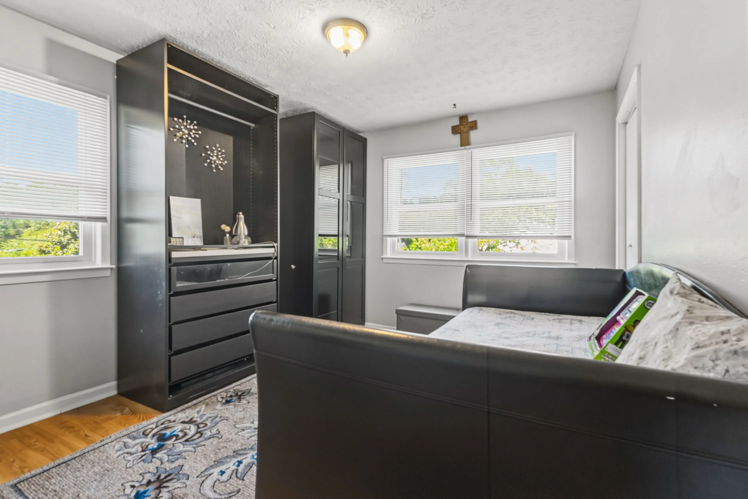 A bedroom with two large windows with white blinds, a dark grey wardrobe, a cross on the wall, and a bed with a floral blanket and pillows. There is a rug with a blue and grey pattern on the wooden floor.