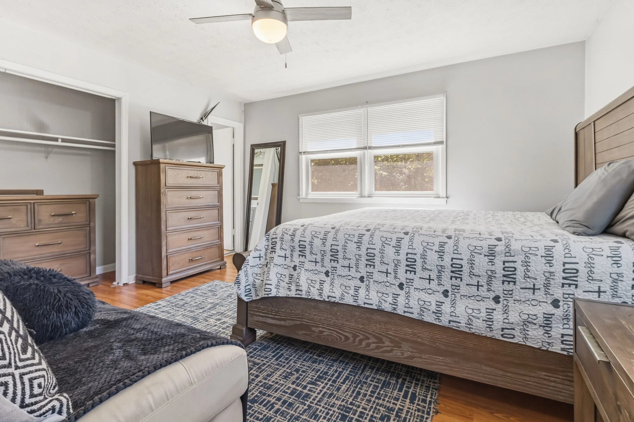 A bedroom with a wooden bed frame, nightstand, dresser, large window with blinds, and a ceiling fan. The bedspread has words like 'LOVE,' 'hope,' and 'blessed.'