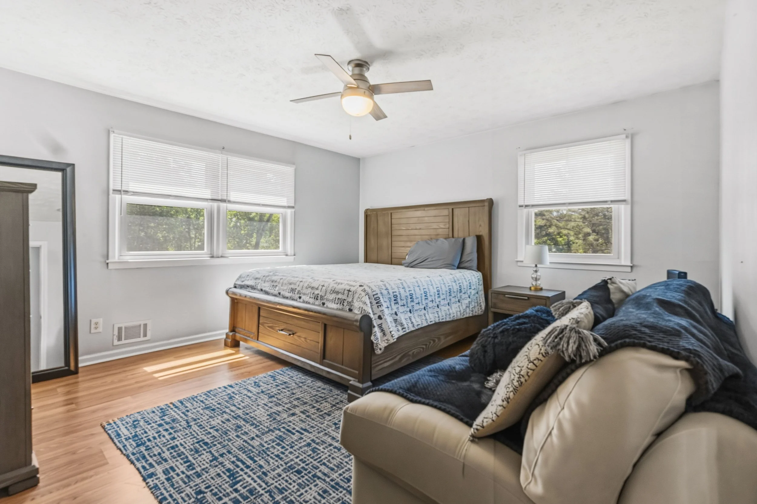 Bedroom with a wooden bed, windows with blinds, a ceiling fan, and a sofa with pillows.