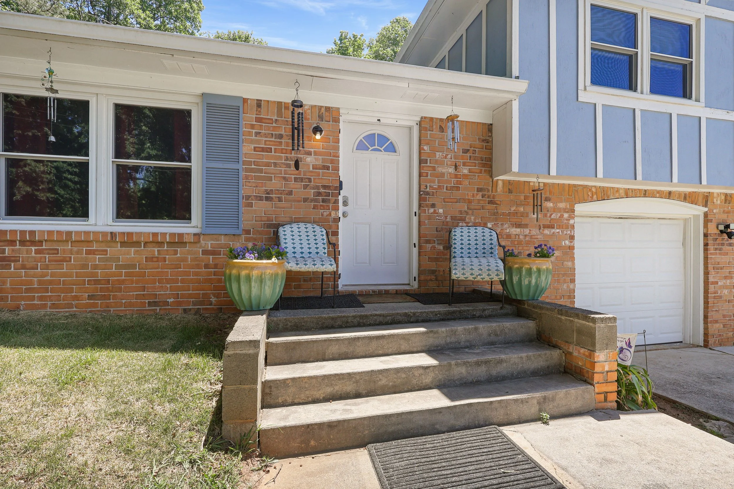 Entrance of a house with brick and light blue siding, concrete steps, two decorative chairs with patterned cushions, large potted plants, wind chimes hanging on the porch, and a white door with a half-moon window.