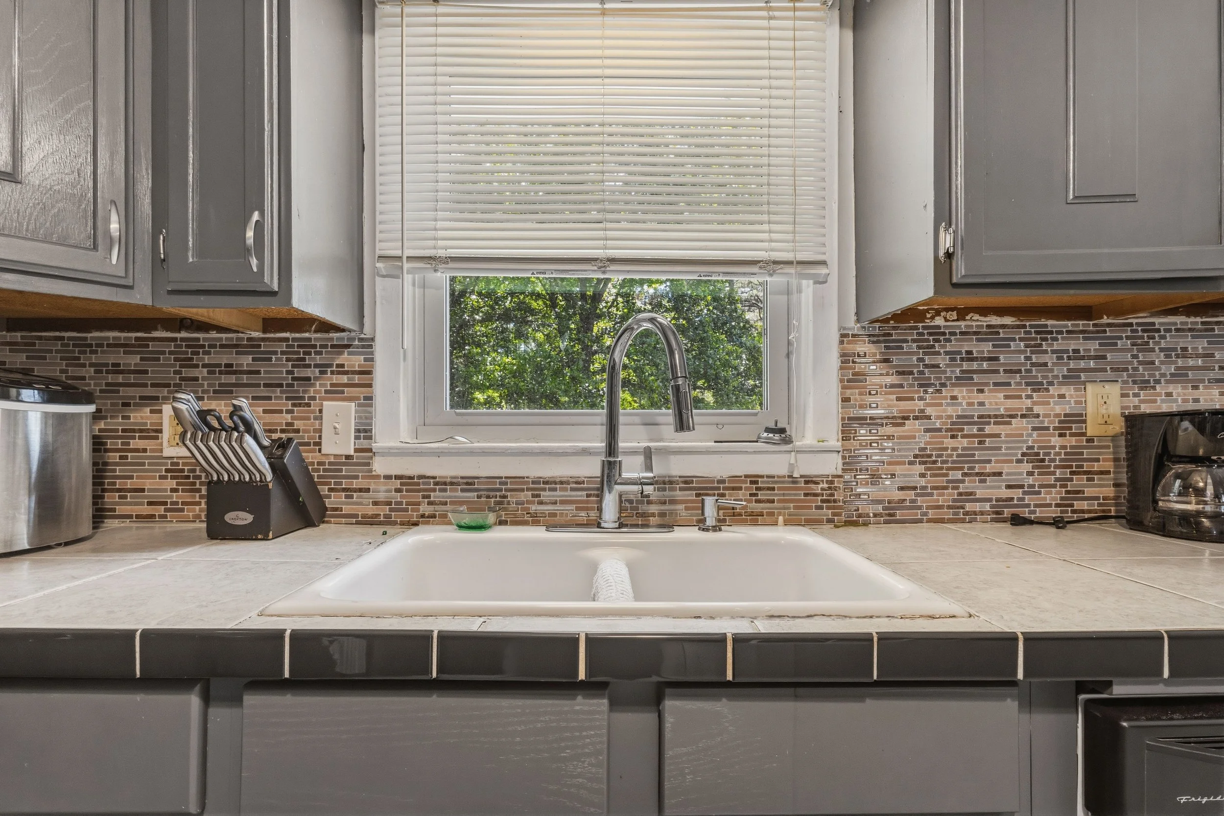 A kitchen sink with a faucet in front of a window with closed white blinds. There is a knife set, stove, and other appliances on the kitchen countertop. The backsplash is made of small, rectangular bricks.