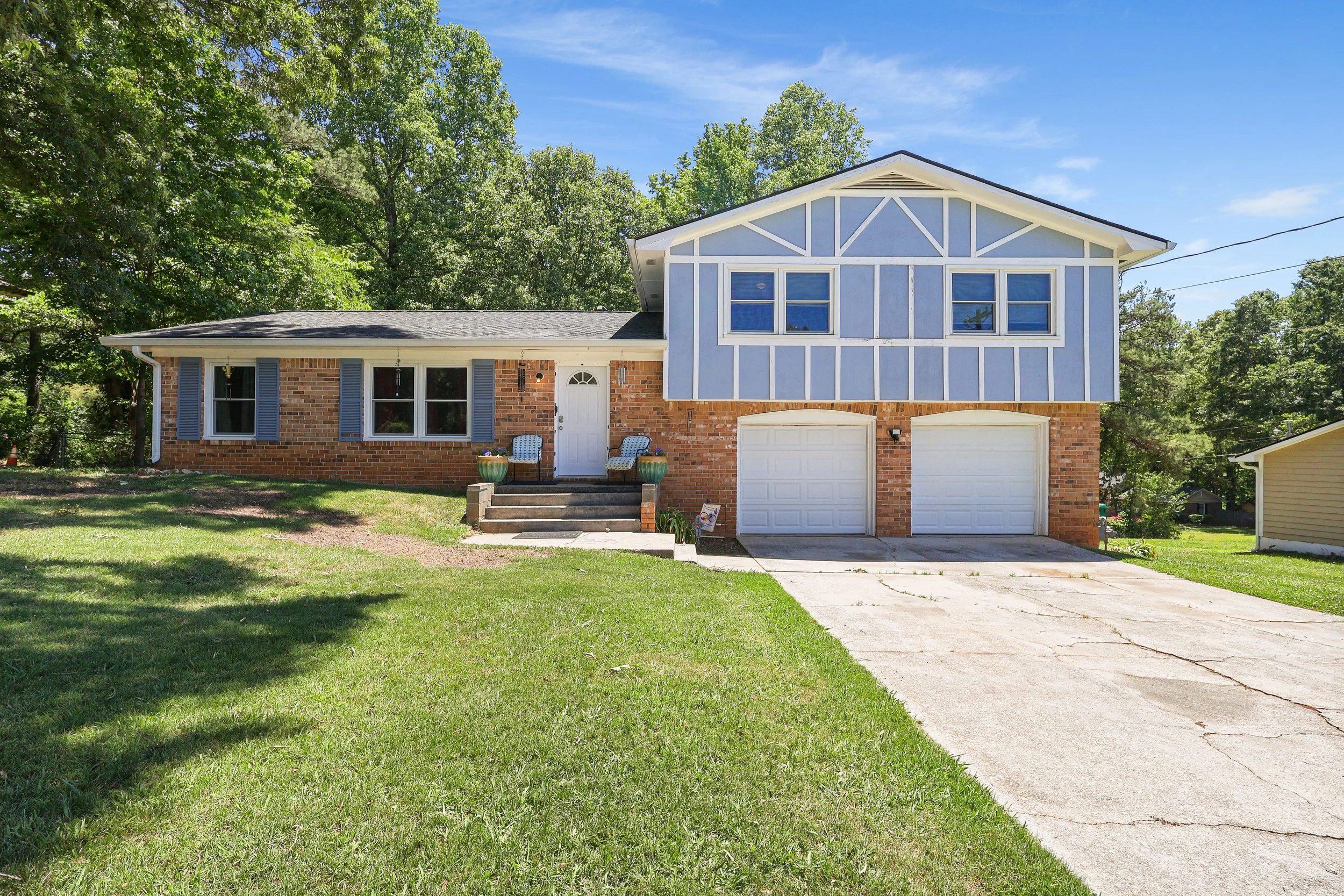 A two-story house with a red brick lower level and a light blue upper level with white trim. The house has two garage doors, a white front door with stairs and potted plants, and windows with blue shutters. The yard has green grass and trees in the b