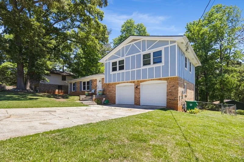Two-story house with brick lower level and blue upper level, white trim, and attached two-car garage, with a concrete driveway and green lawn, surrounded by trees.