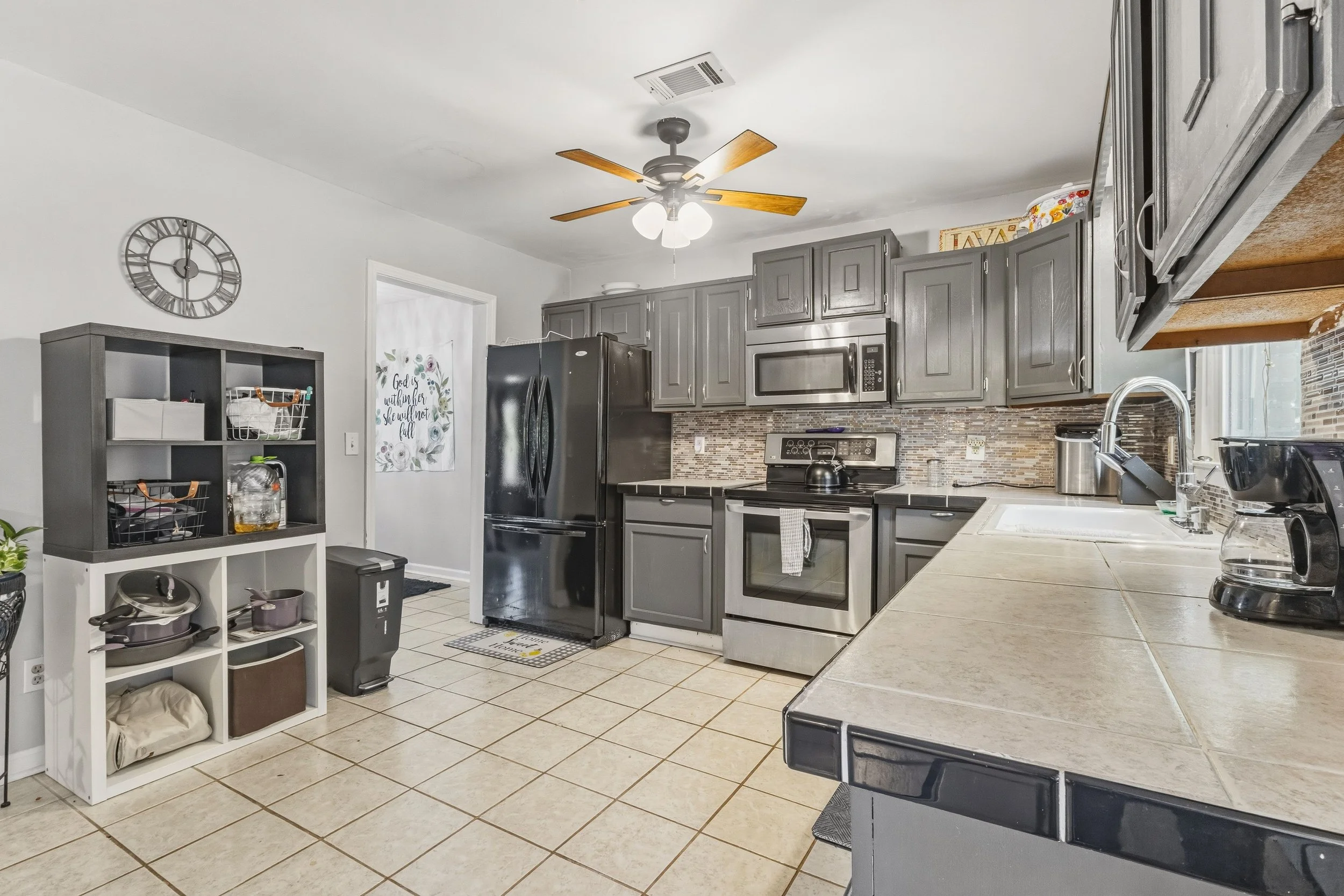 A kitchen with gray cabinets, a black refrigerator, and a tiled backsplash. There is a ceiling fan with wooden blades and light fixtures, a gray shelving unit, and various kitchen appliances on the countertops.