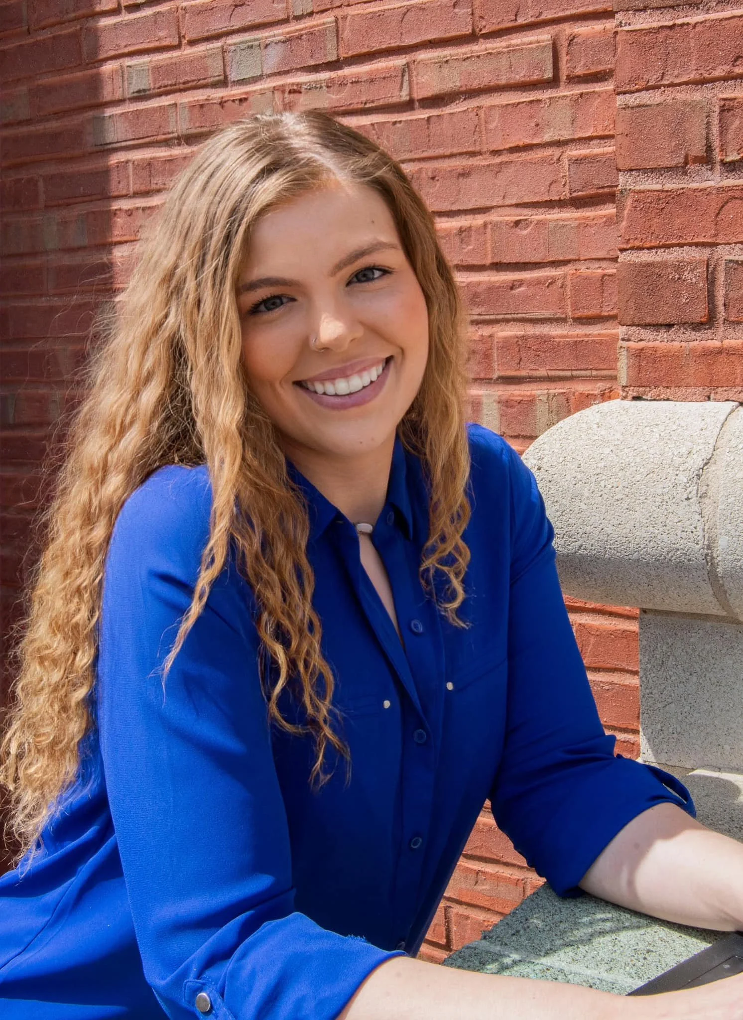 A woman with long curly blonde hair, wearing a blue shirt, smiling and sitting against a red brick wall.