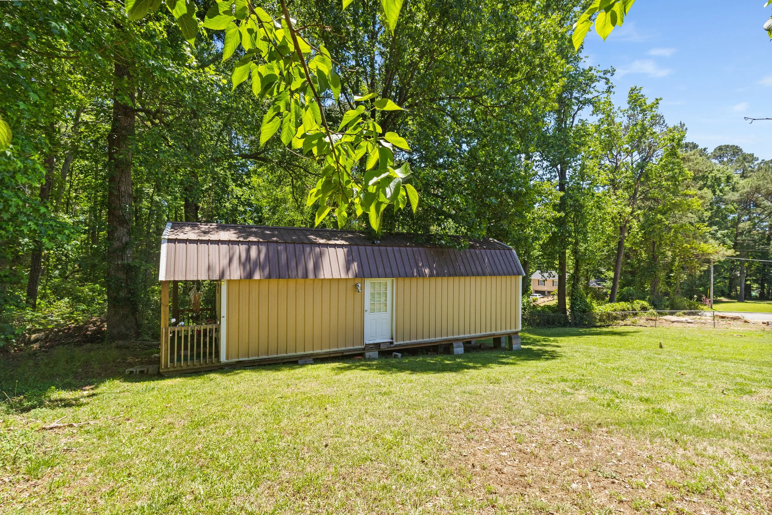 A small yellow shed with a metal roof situated in a grassy backyard surrounded by trees under a blue sky.