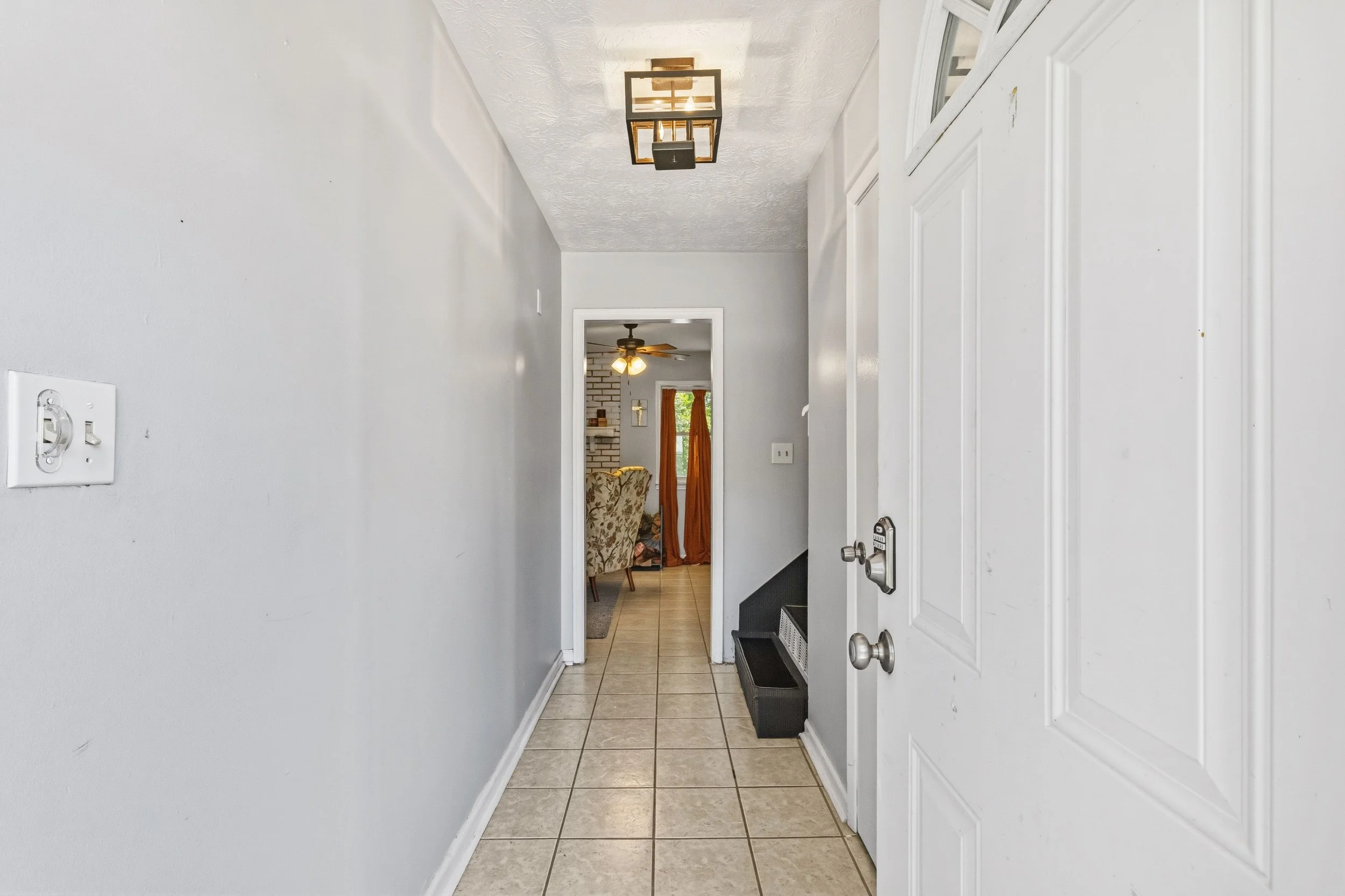A hallway with white walls and tiled flooring leading to a living room area with a ceiling fan, fireplace, and window with orange curtains.