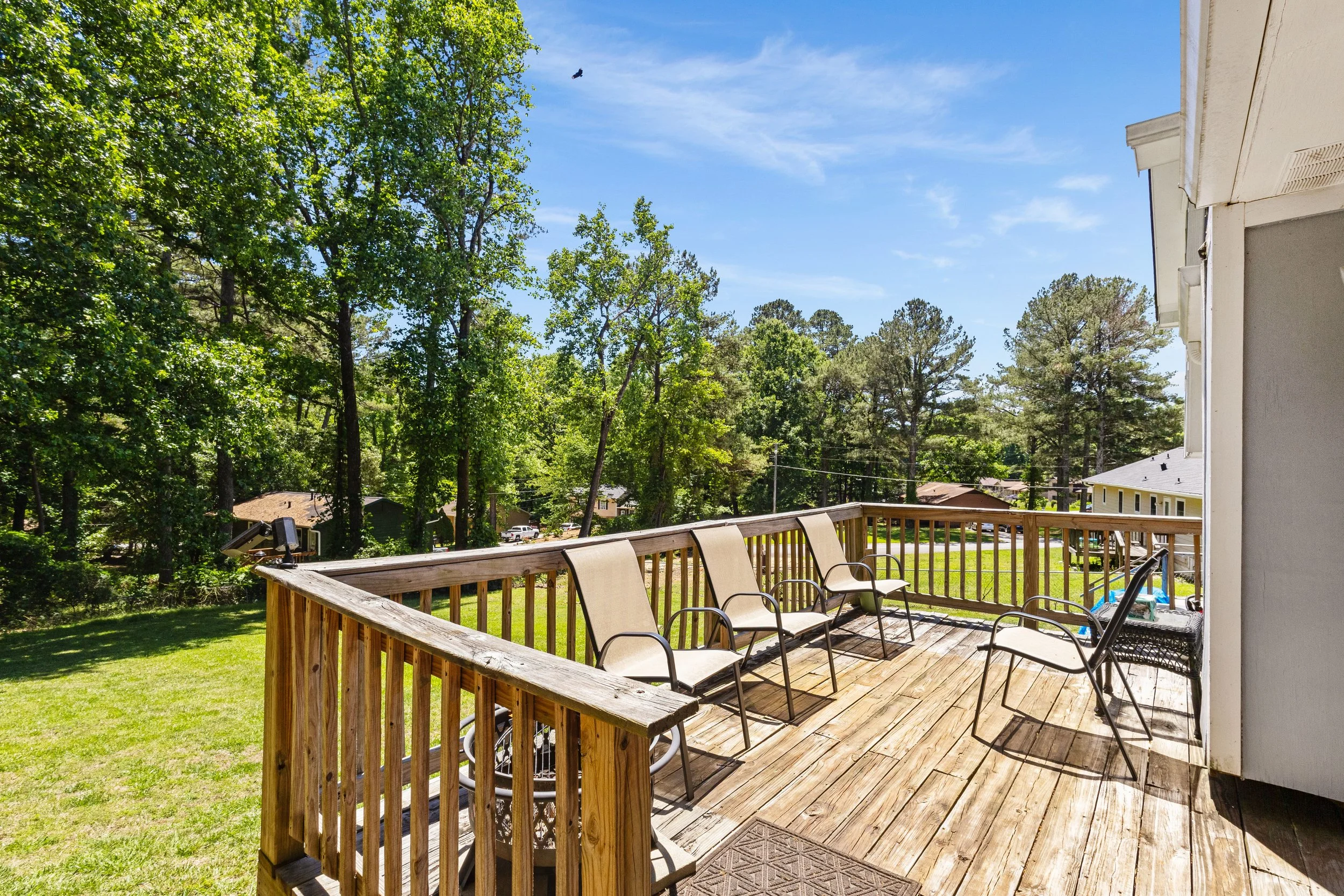 A wooden deck with outdoor chairs overlooking a yard with lush green trees under a blue sky with a few clouds.
