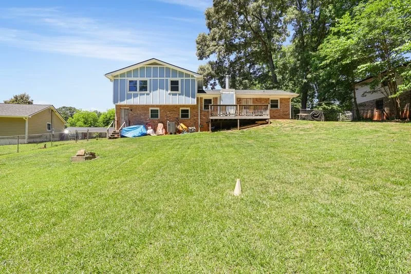 Backyard with green grass, a house with a brick and blue exterior, a wooden deck, some outdoor items covered with a tarp, and large trees in the background under a clear blue sky.