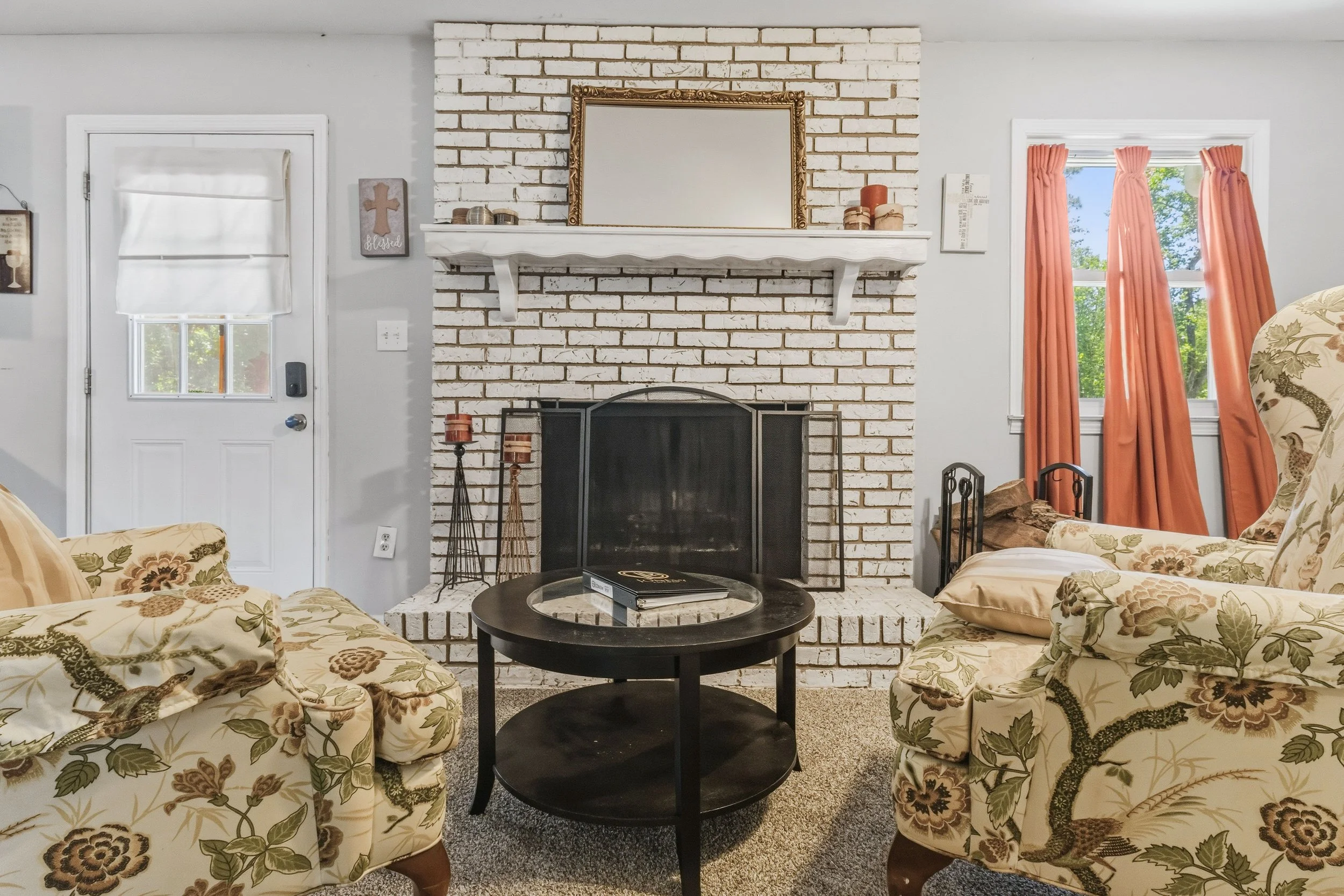 Living room with white brick fireplace, framed mirror, floral patterned sofas, black coffee table, and windows with orange curtains.