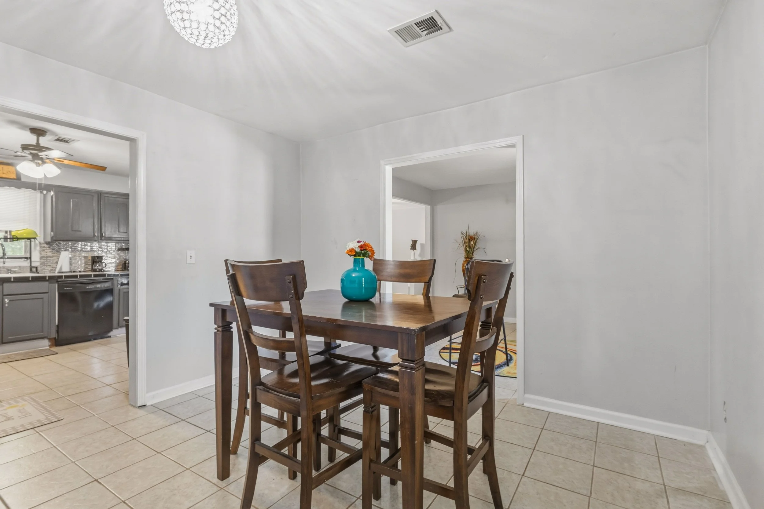 A dining room with a wooden table and six matching chairs. A teal vase with orange and yellow flowers is on the table. The room has beige tiled floors and white walls. Visible in the background is a doorway leading to a kitchen with gray cabinets, a 